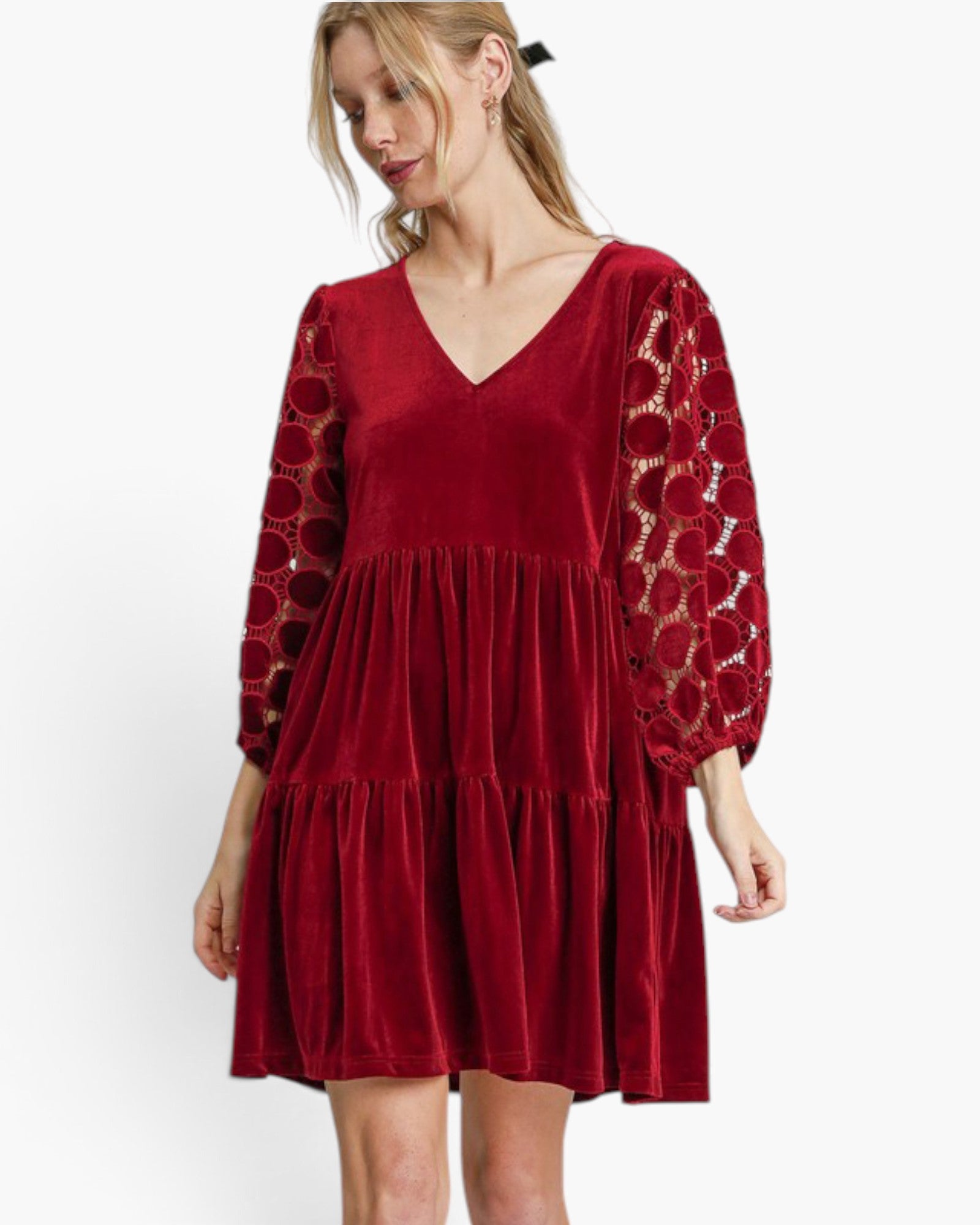 A woman wearing a short red velvet dress with sheer patterned lace sleeves, photographed against a plain white studio background.