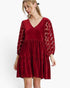 A woman wearing a short red velvet dress with sheer patterned lace sleeves, photographed against a plain white studio background.