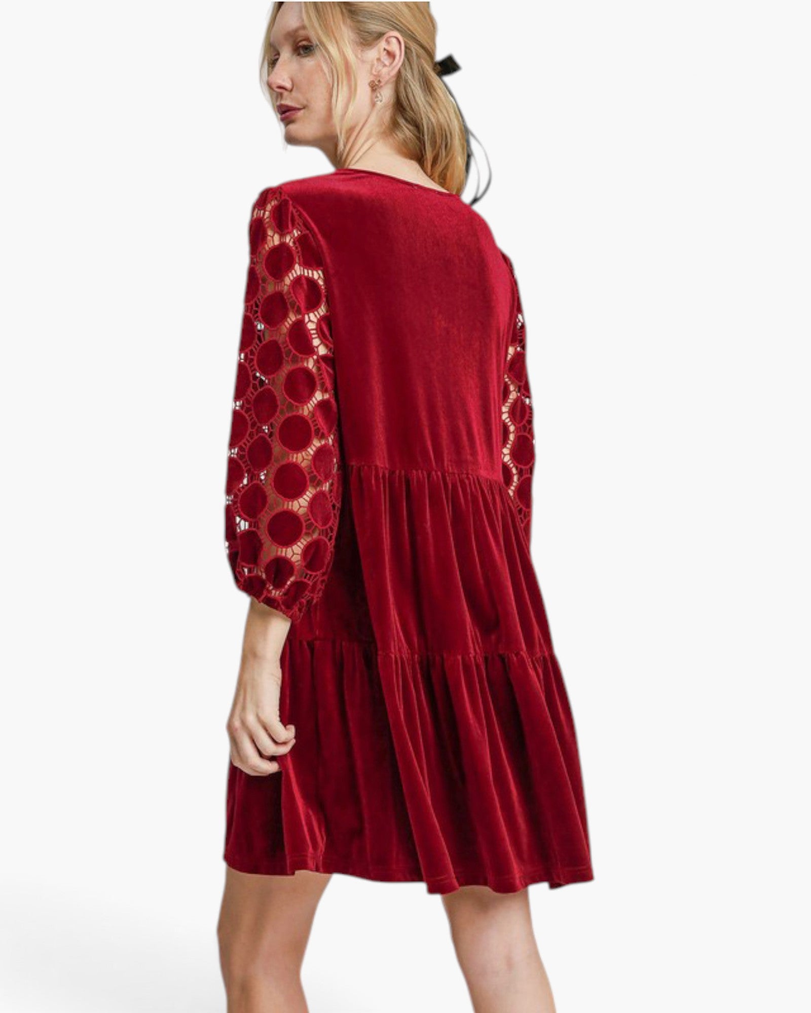A woman modeling a red velvet tiered dress with sheer patterned sleeves, shown from a three-quarter back angle against a plain white studio background.