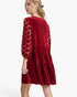 A woman modeling a red velvet tiered dress with sheer patterned sleeves, shown from a three-quarter back angle against a plain white studio background.