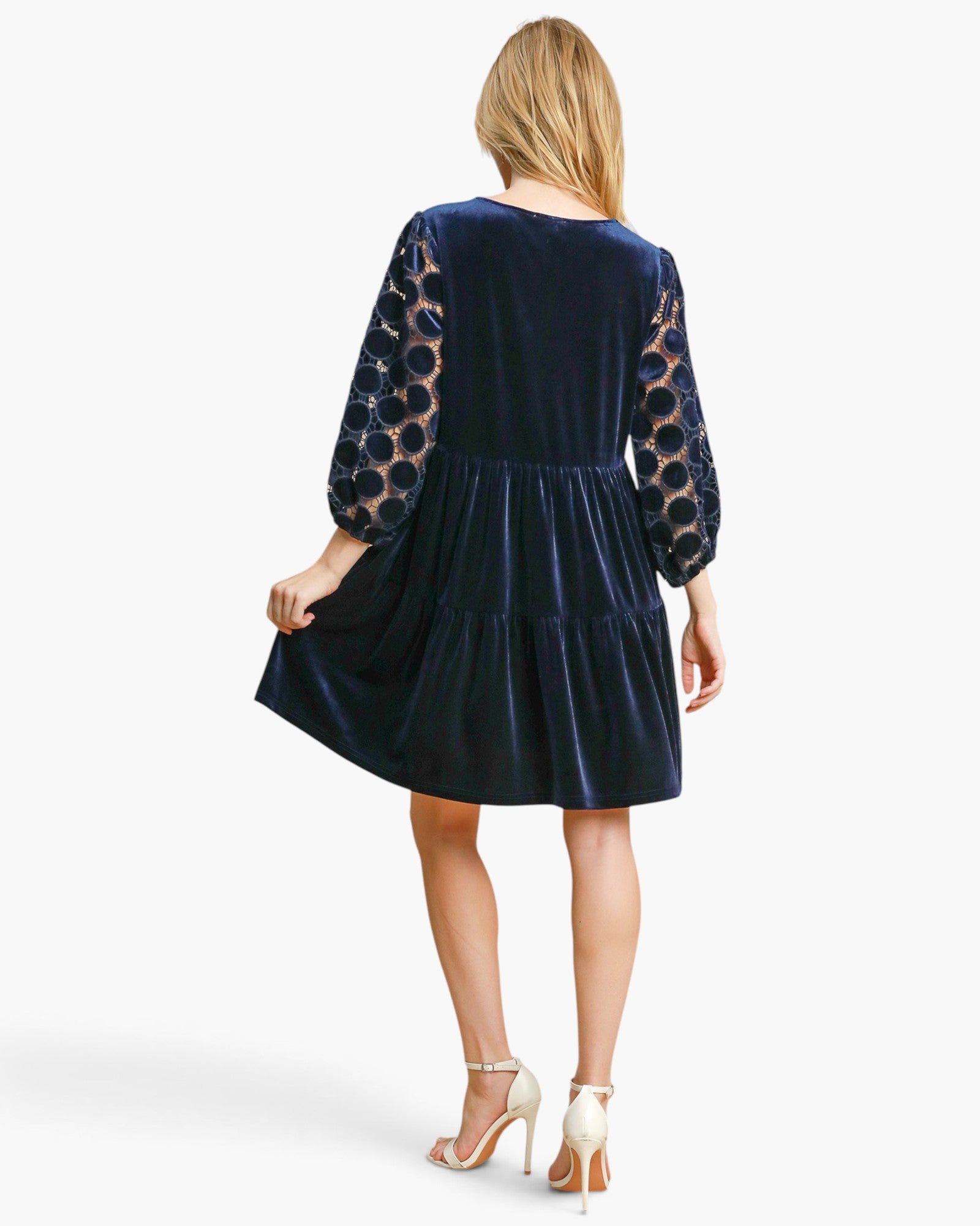 A woman shown from the back wearing a navy blue velvet dress with sheer patterned sleeves, paired with white high-heeled sandals, against a light gray studio background.