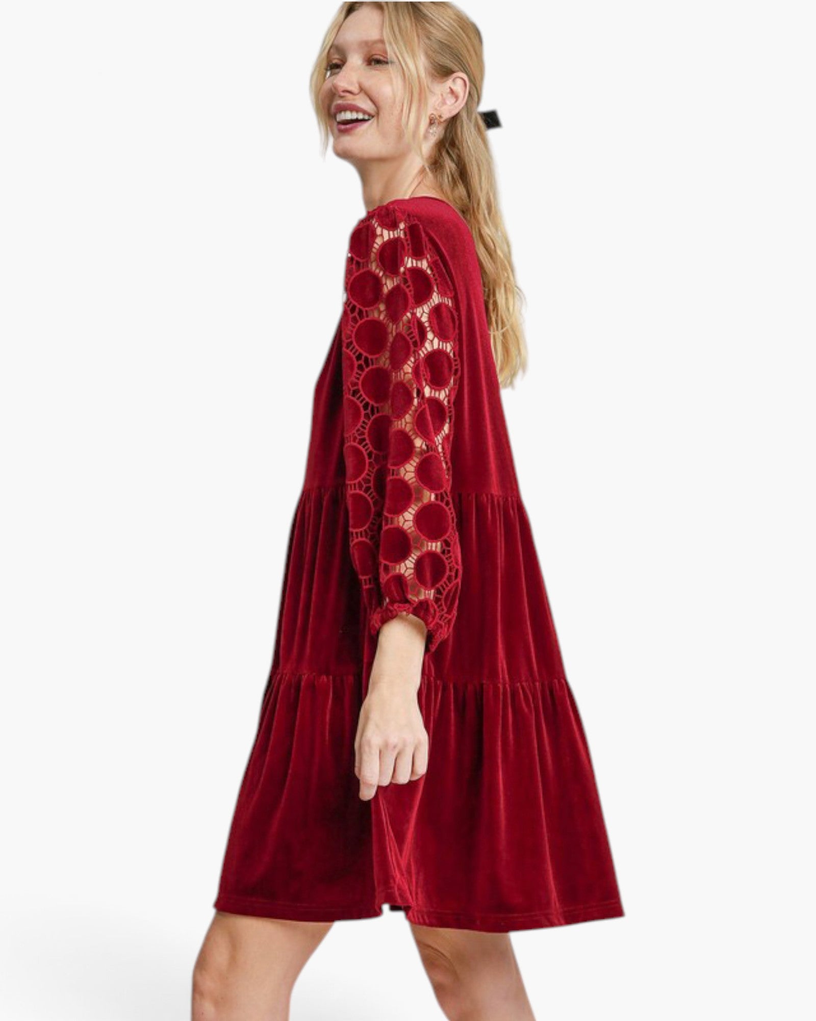 A woman wearing a red velvet tiered dress with sheer floral lace long sleeves, shown from a side-back angle against a plain white studio background.