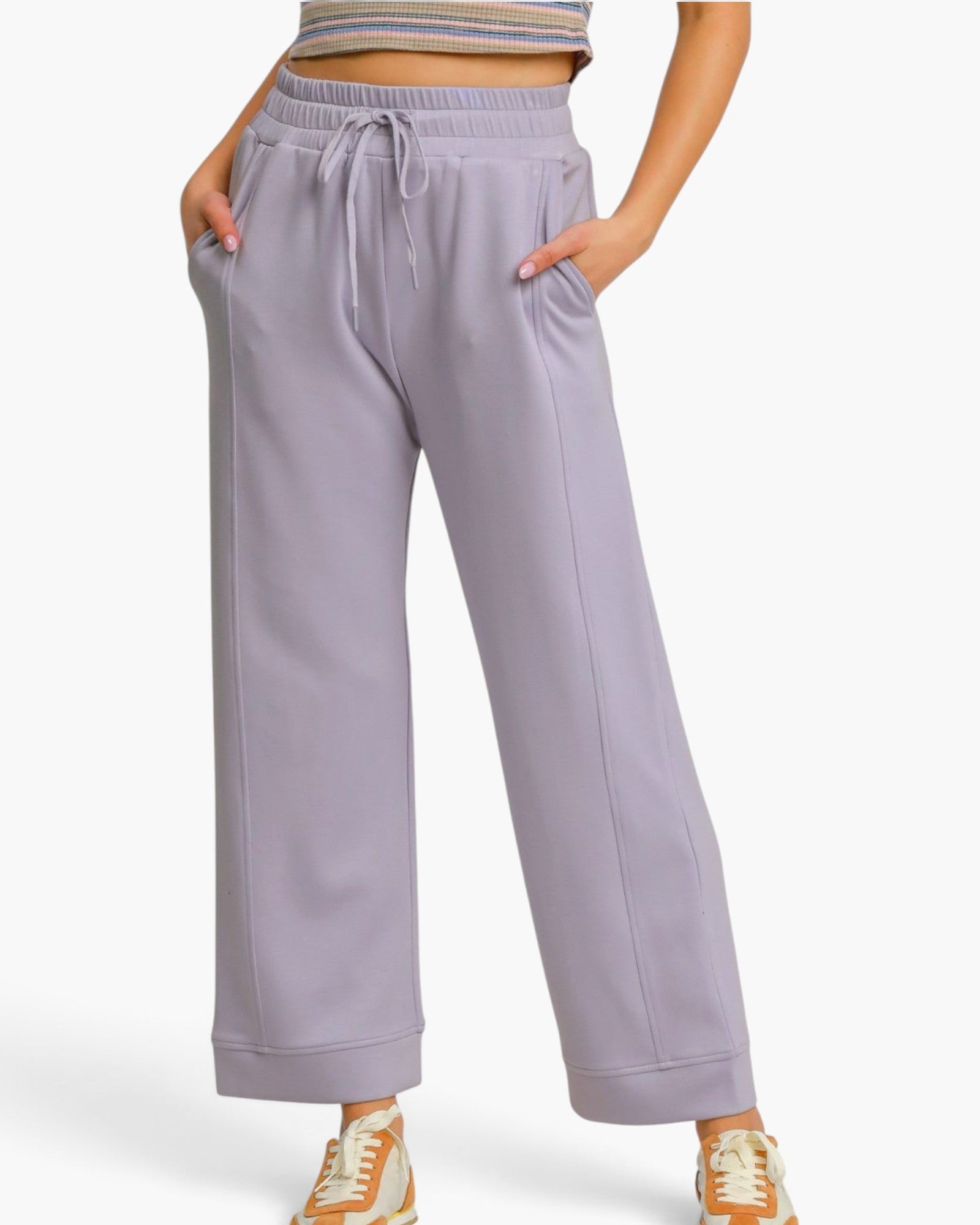 A person wearing light lavender wide-leg sweatpants with a drawstring waistband and side pockets, paired with a striped crop top and beige-and-white sneakers, against a plain light gray studio background.