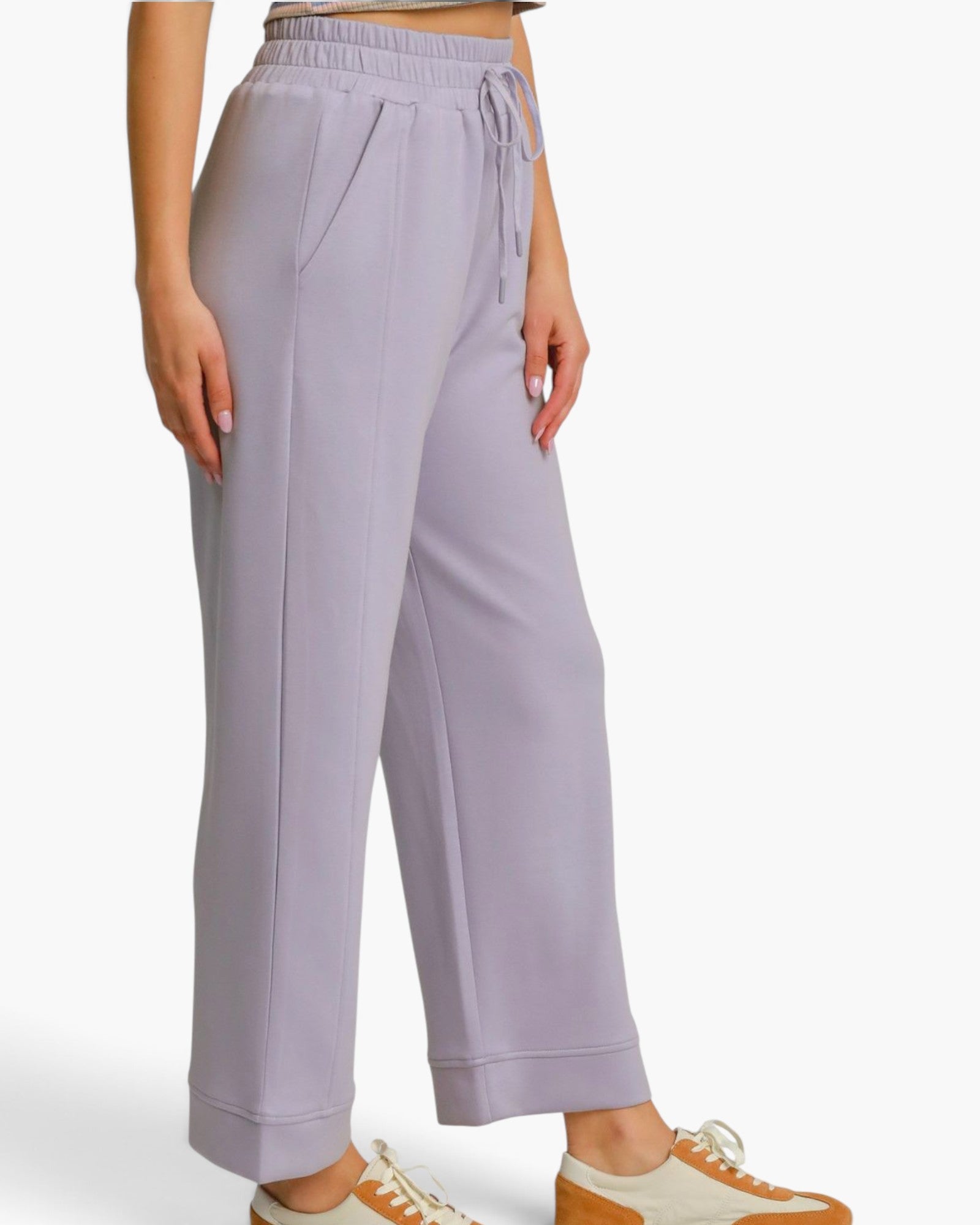 A person wearing light lavender wide-leg sweatpants with an elastic drawstring waist, paired with white and tan sneakers, shown against a plain light gray studio background.