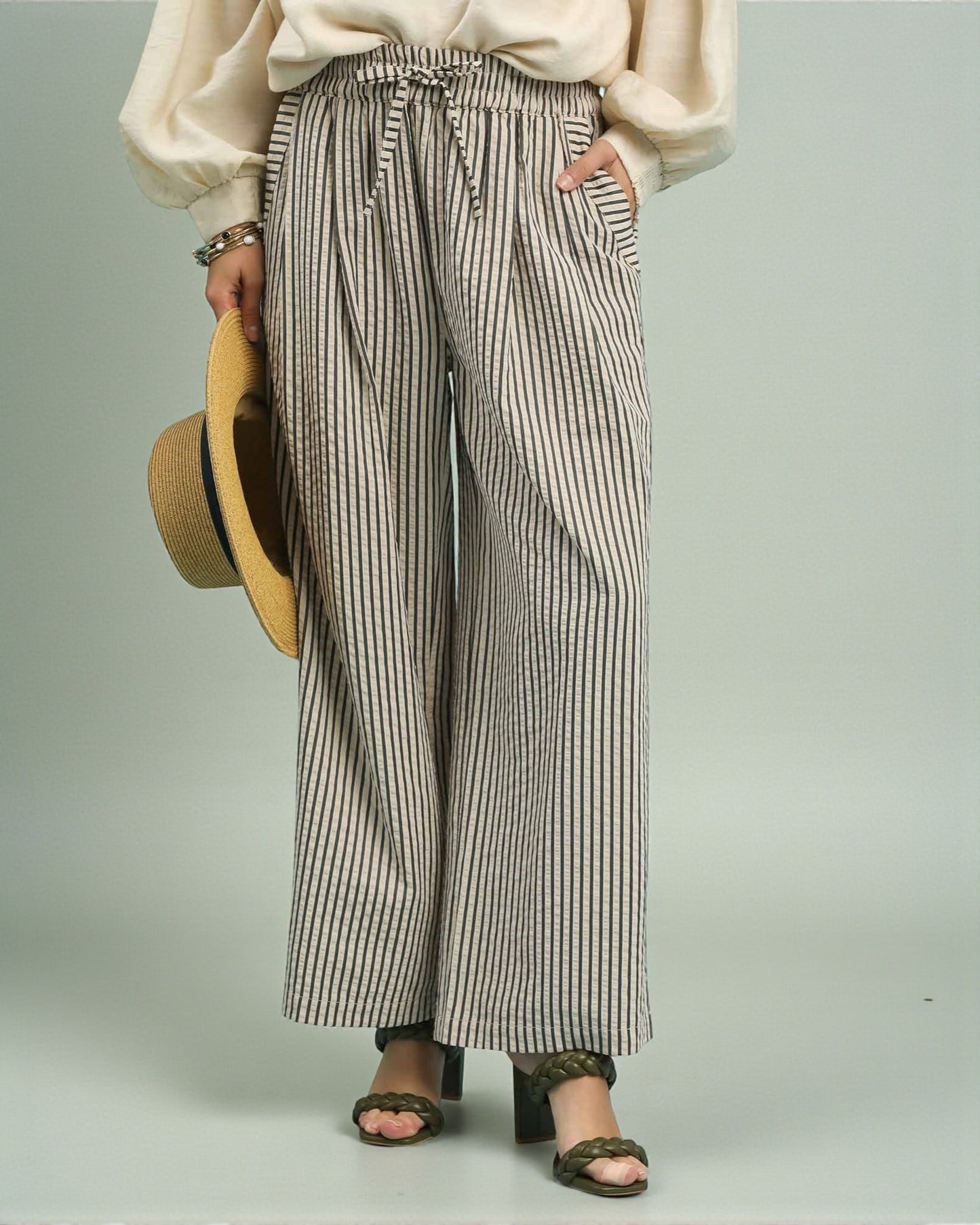 Person wearing striped wide-leg pants holding a straw hat on a plain background