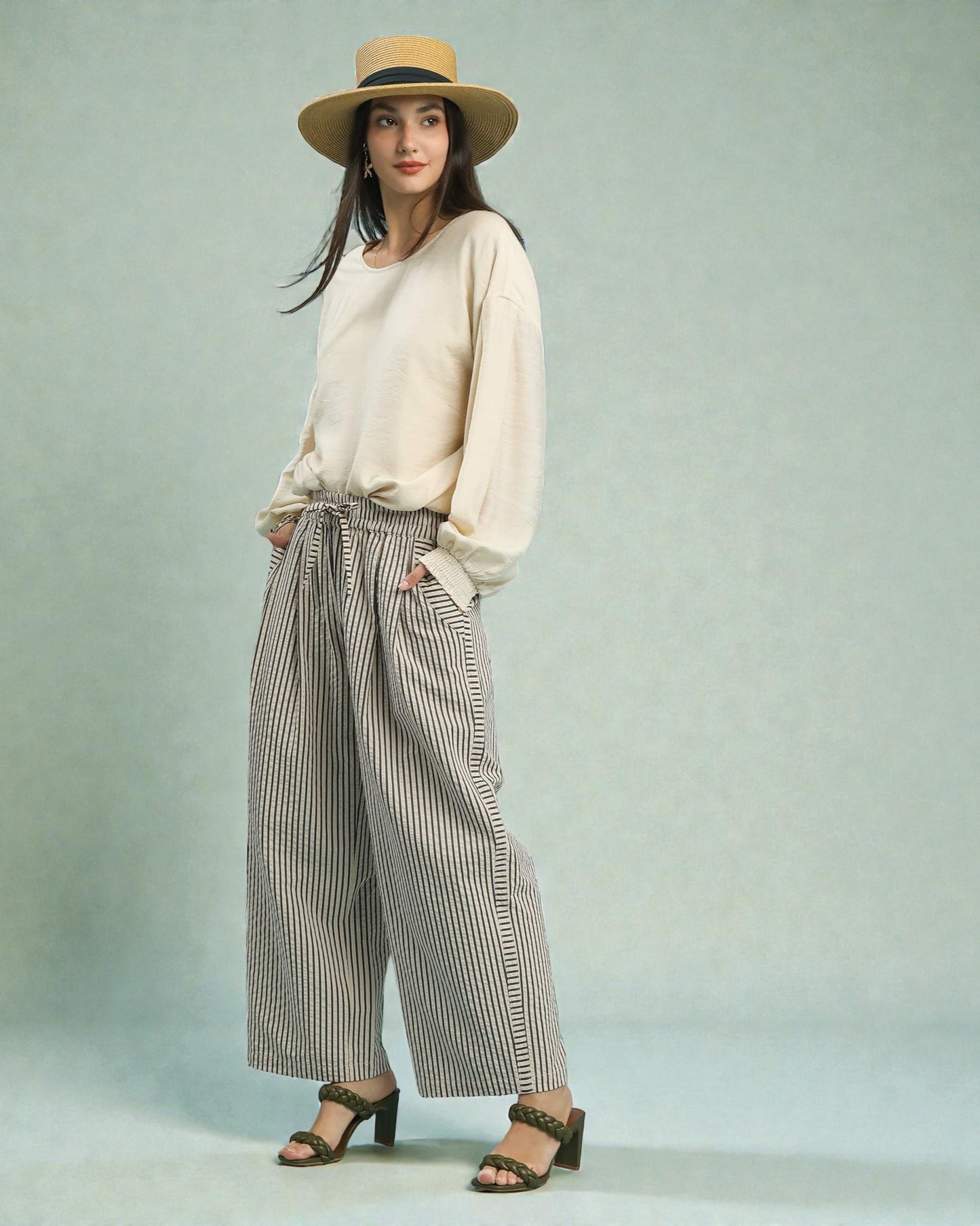 Woman wearing a beige sweater, striped pants, and a straw hat on a light blue background