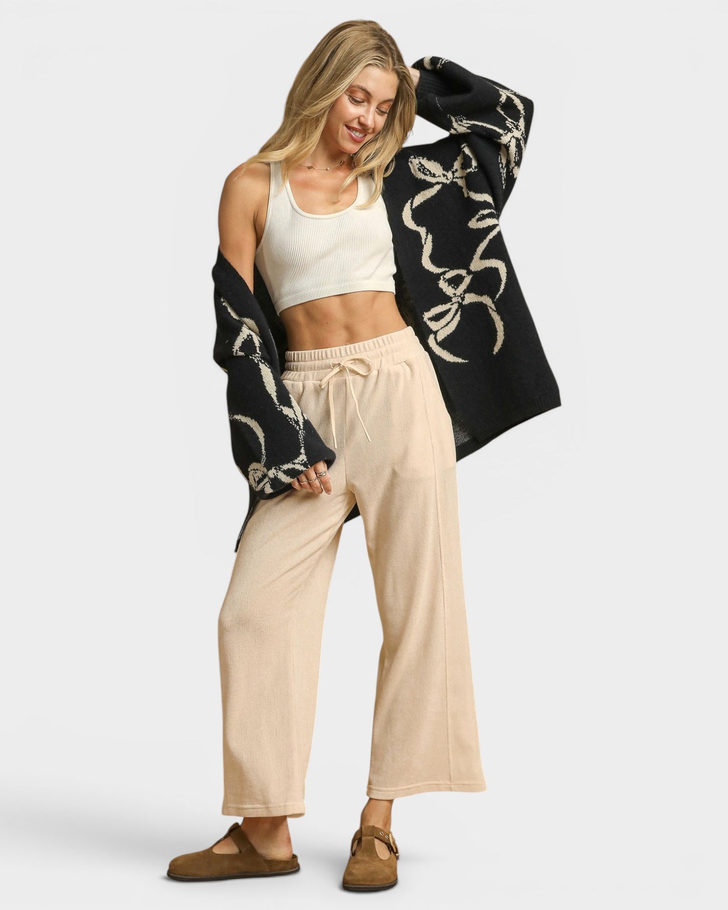 Woman wearing a black patterned cardigan, white top, beige pants, and brown shoes on a white background