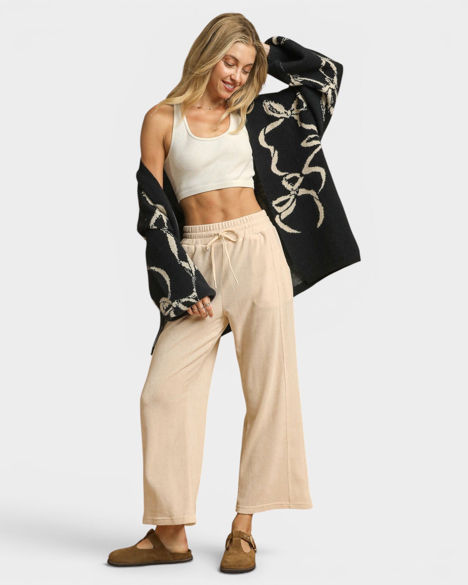Woman wearing a black patterned cardigan, white top, beige pants, and brown shoes on a white background