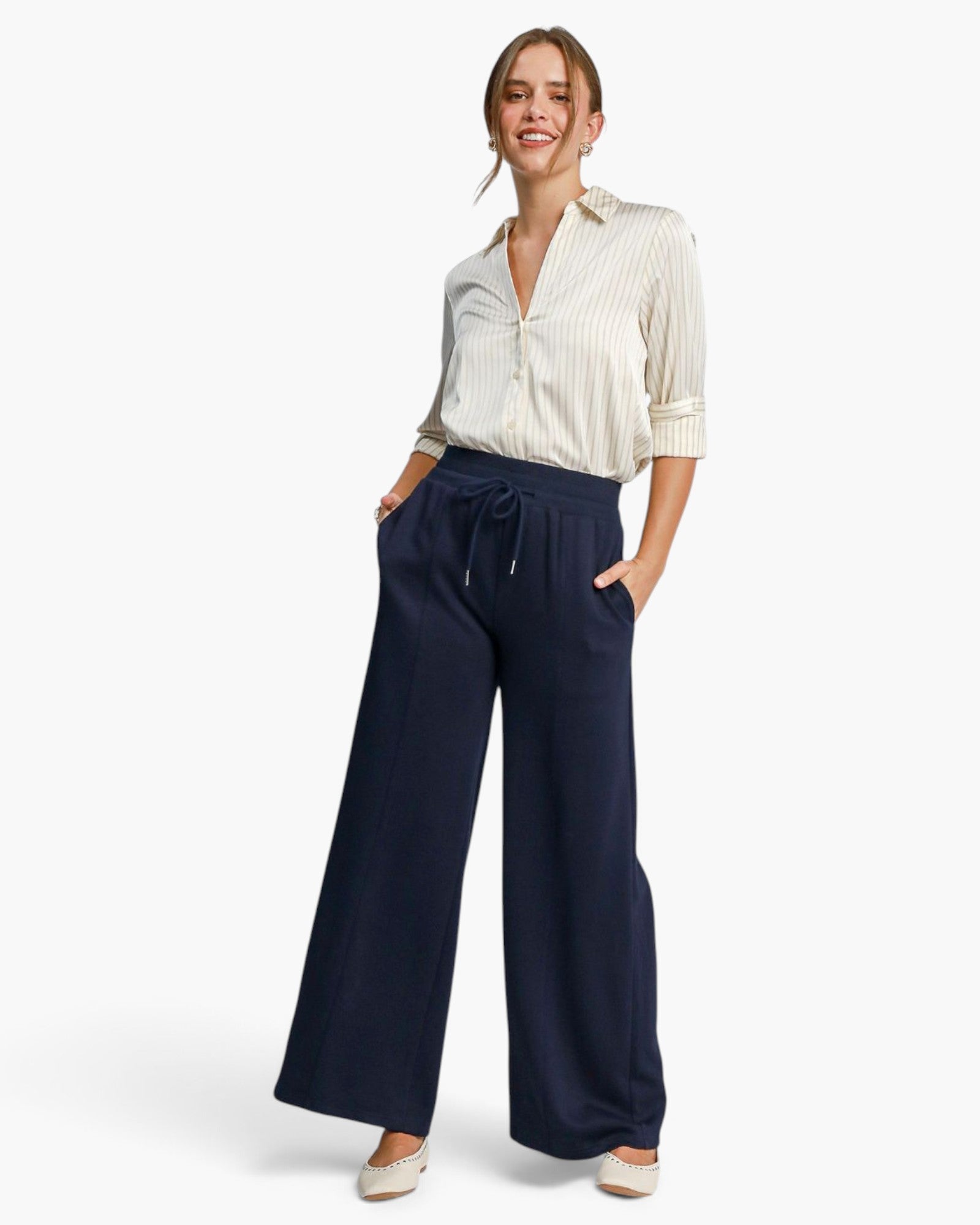 A woman modeling a cream and white vertically striped button-down shirt paired with navy blue wide-leg drawstring pants, standing against a clean white studio background.