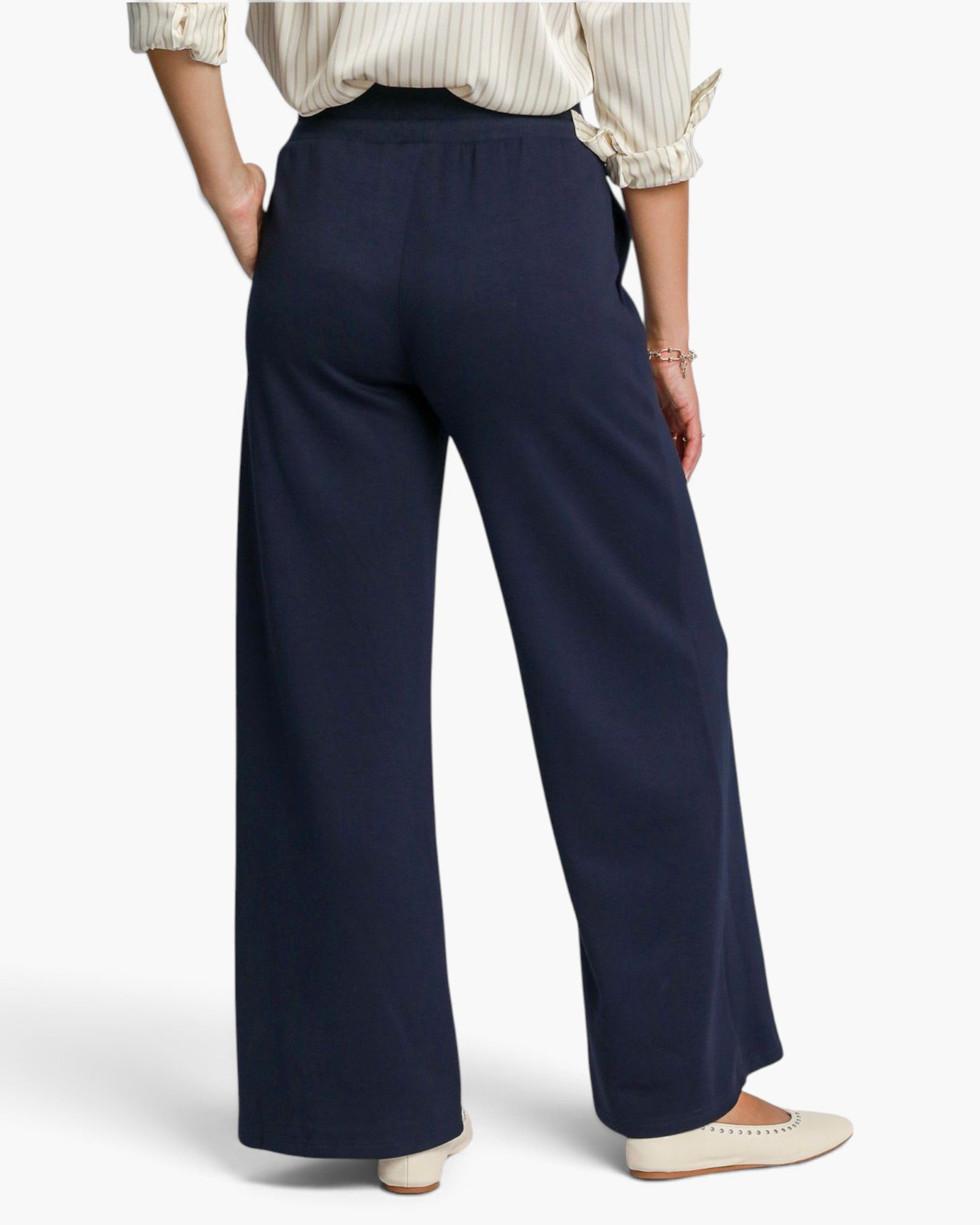 A model shown from the back wearing navy blue wide-leg trousers, a cream and white striped blouse, and cream flat shoes against a light gray studio background.
