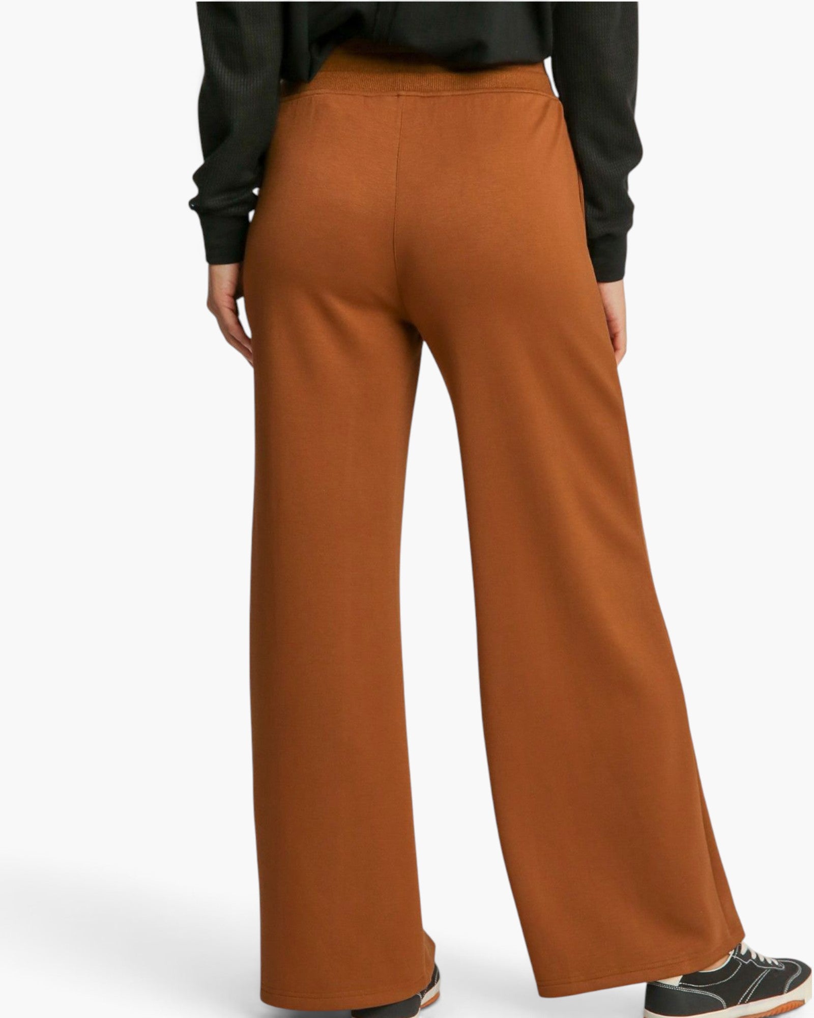 A person shown from the back wearing wide-leg rust-colored pants, a black long-sleeve top, and black sneakers against a clean white studio background.