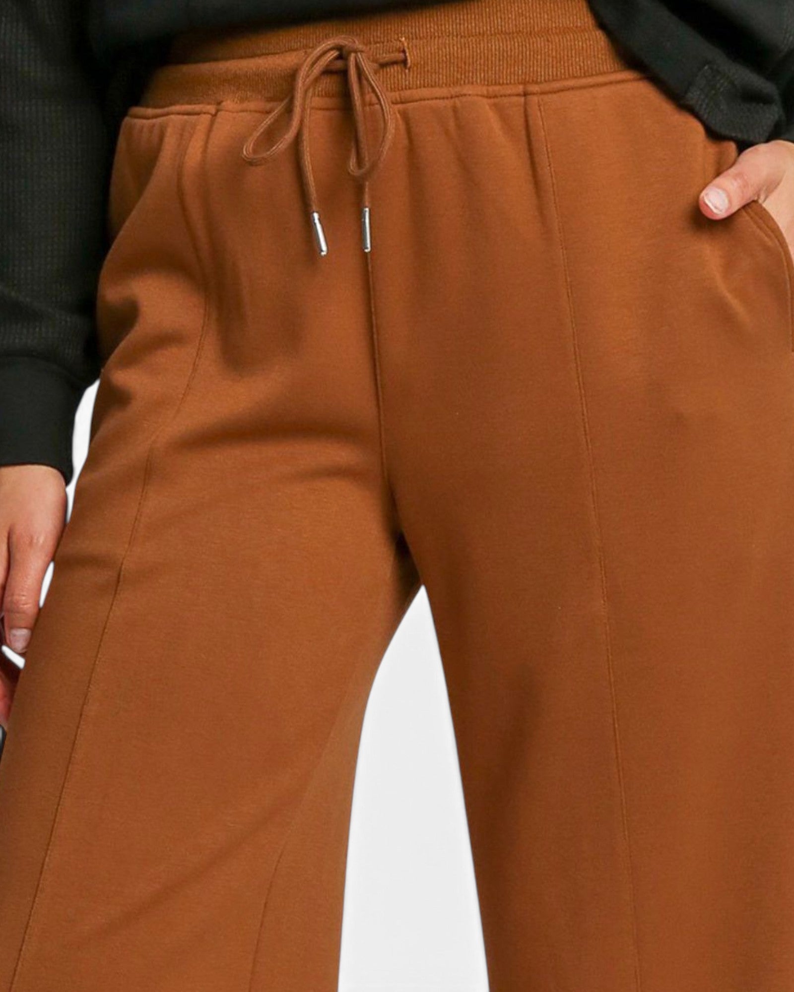 Close-up of a person wearing rust-brown drawstring sweatpants with a black top, showing the waistband, drawstrings, side pocket, and hand.