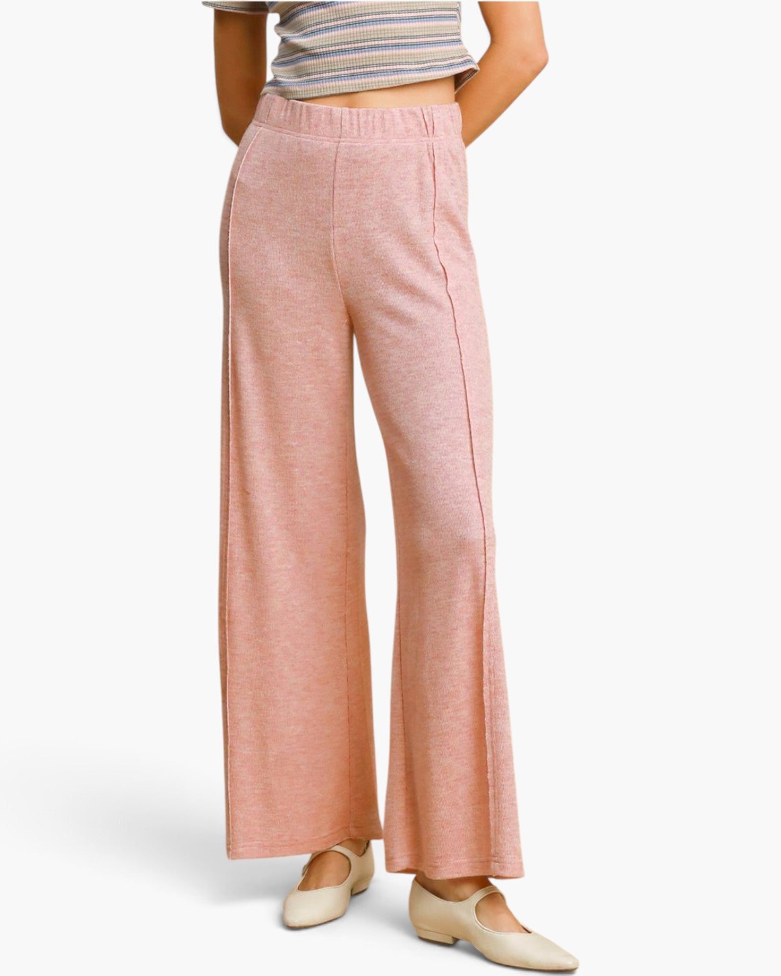 A model wearing a pair of dusty pink wide-leg knit pants, paired with a striped crop top and beige flats, shown from the waist down against a light gray studio background.