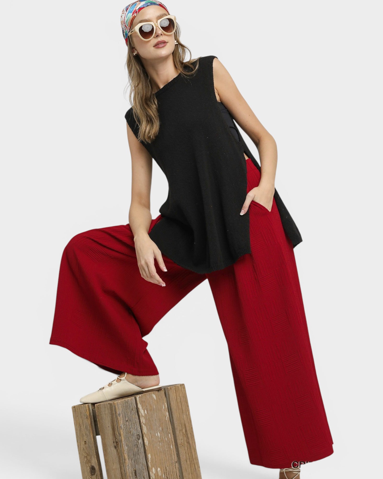 Woman wearing a black sleeveless top and red pants sitting on a wooden crate against a white background.