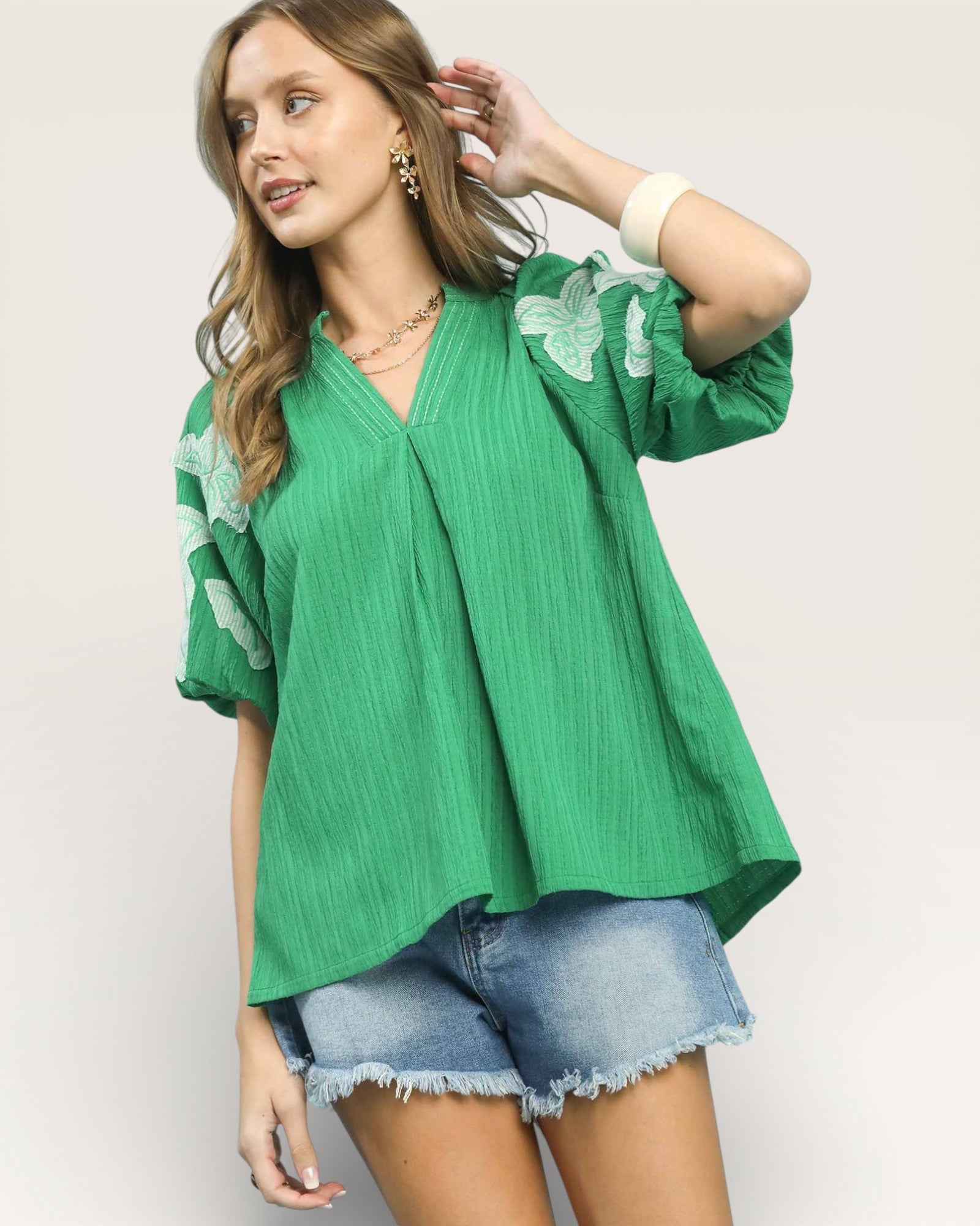 Woman wearing a green blouse with floral sleeves and denim shorts on a plain background