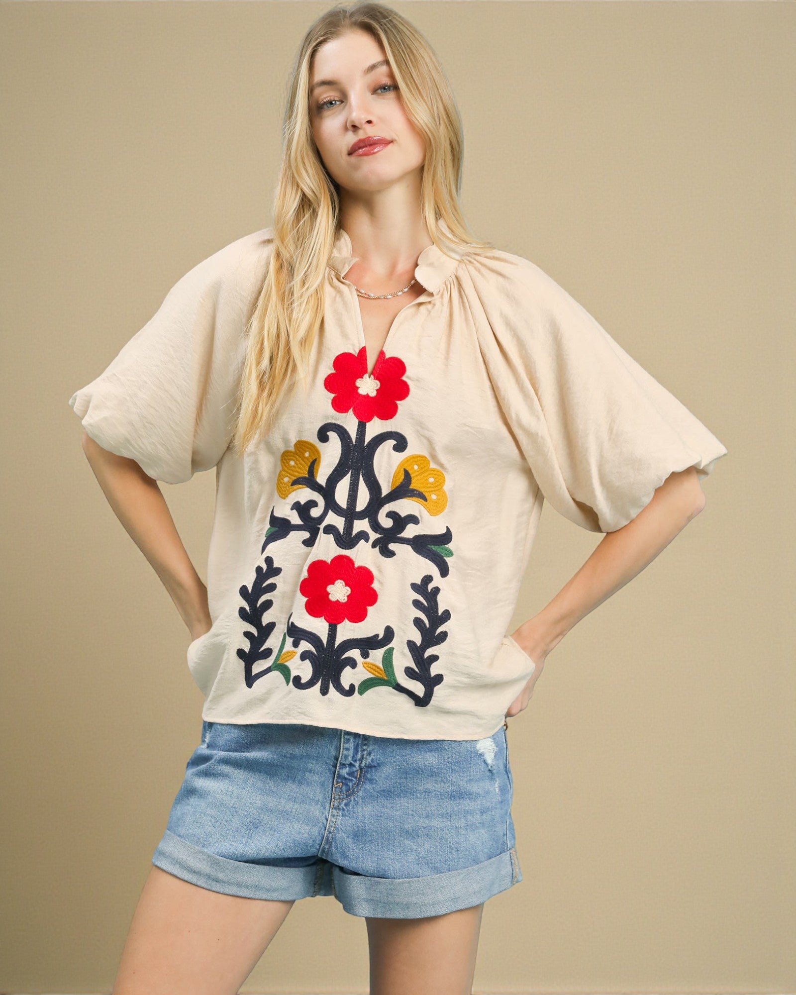 Woman wearing a beige blouse with floral embroidery and denim shorts on a beige background