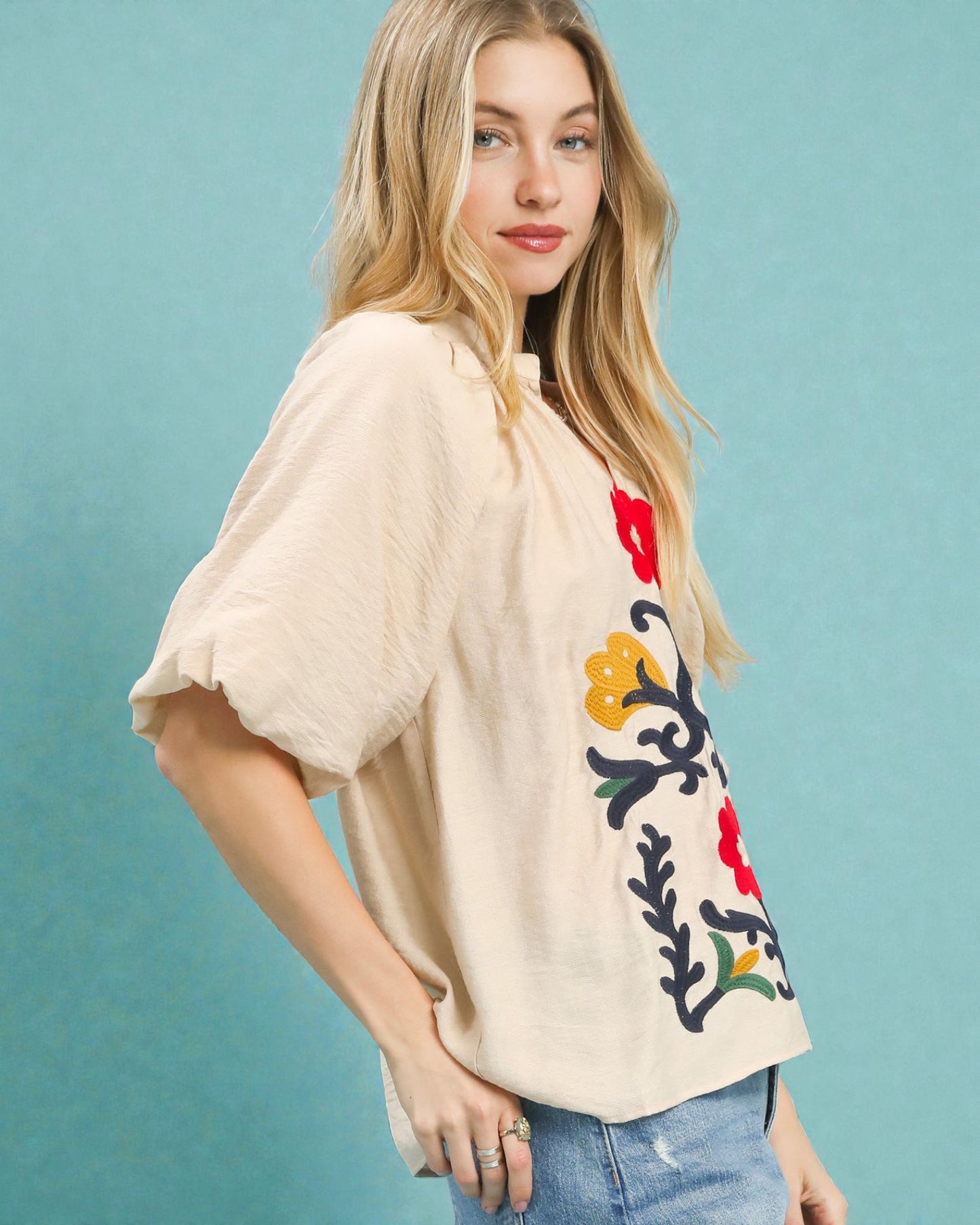 Woman wearing a beige blouse with floral embroidery against a teal background