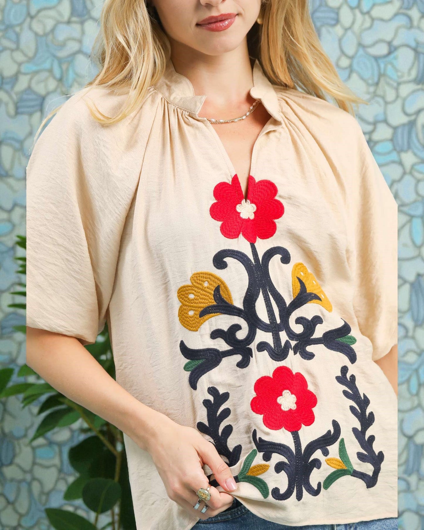 Woman wearing a beige top with floral embroidery against a textured wall.