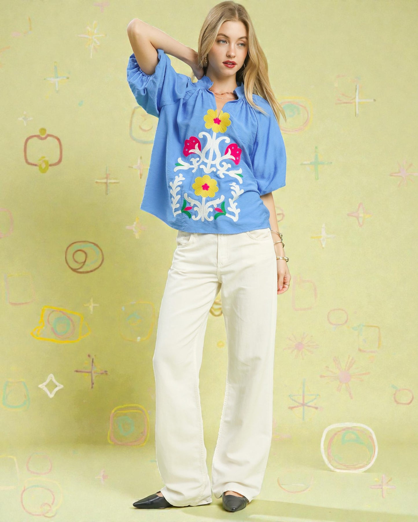 Woman wearing a blue blouse with floral embroidery and white pants on a light green background