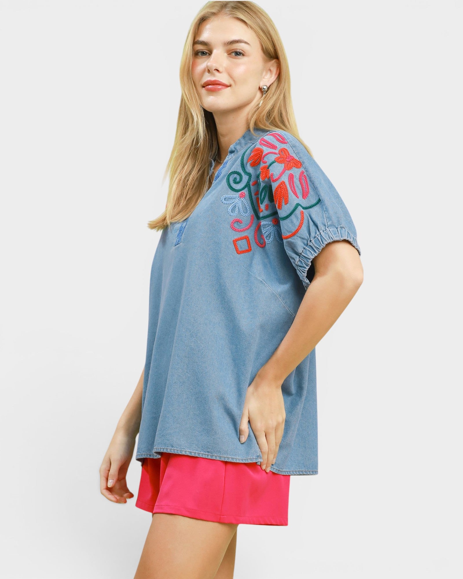Woman wearing a blue denim top with colorful embroidery and pink shorts on a white background
