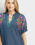 Woman wearing a blue denim blouse with colorful embroidery on a plain background