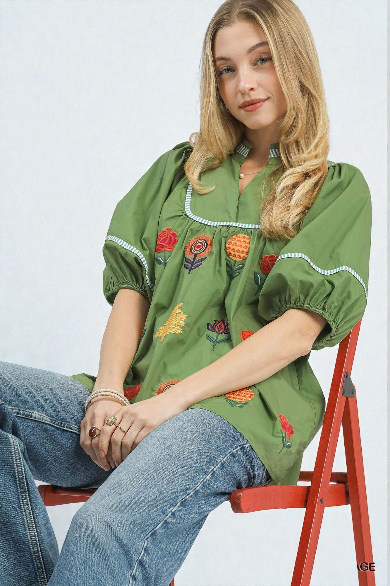 Woman wearing a green embroidered blouse sitting on a red chair against a white background