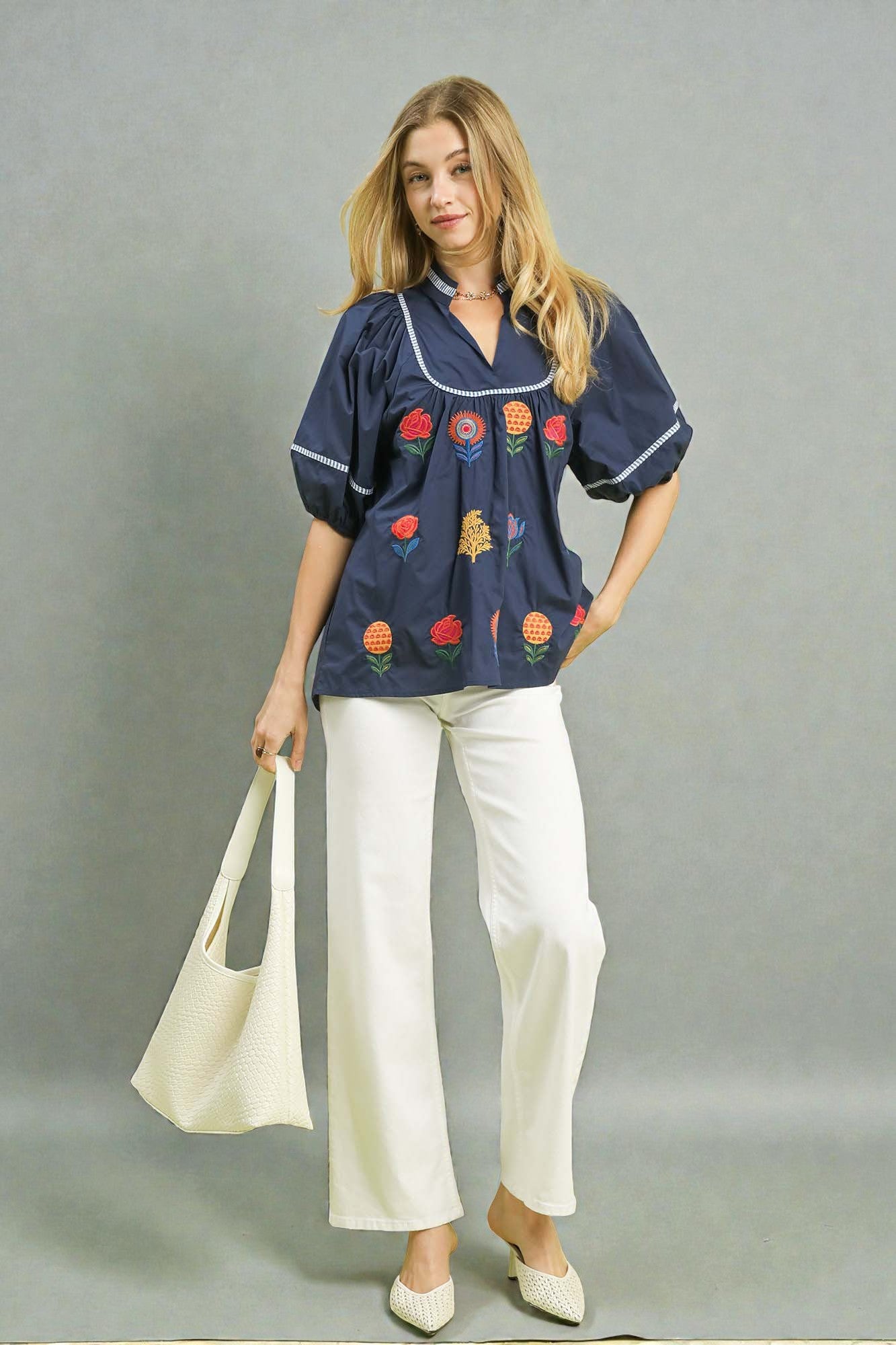 Woman wearing a navy blouse with colorful embroidery and white pants, holding a beige bag on a gray background
