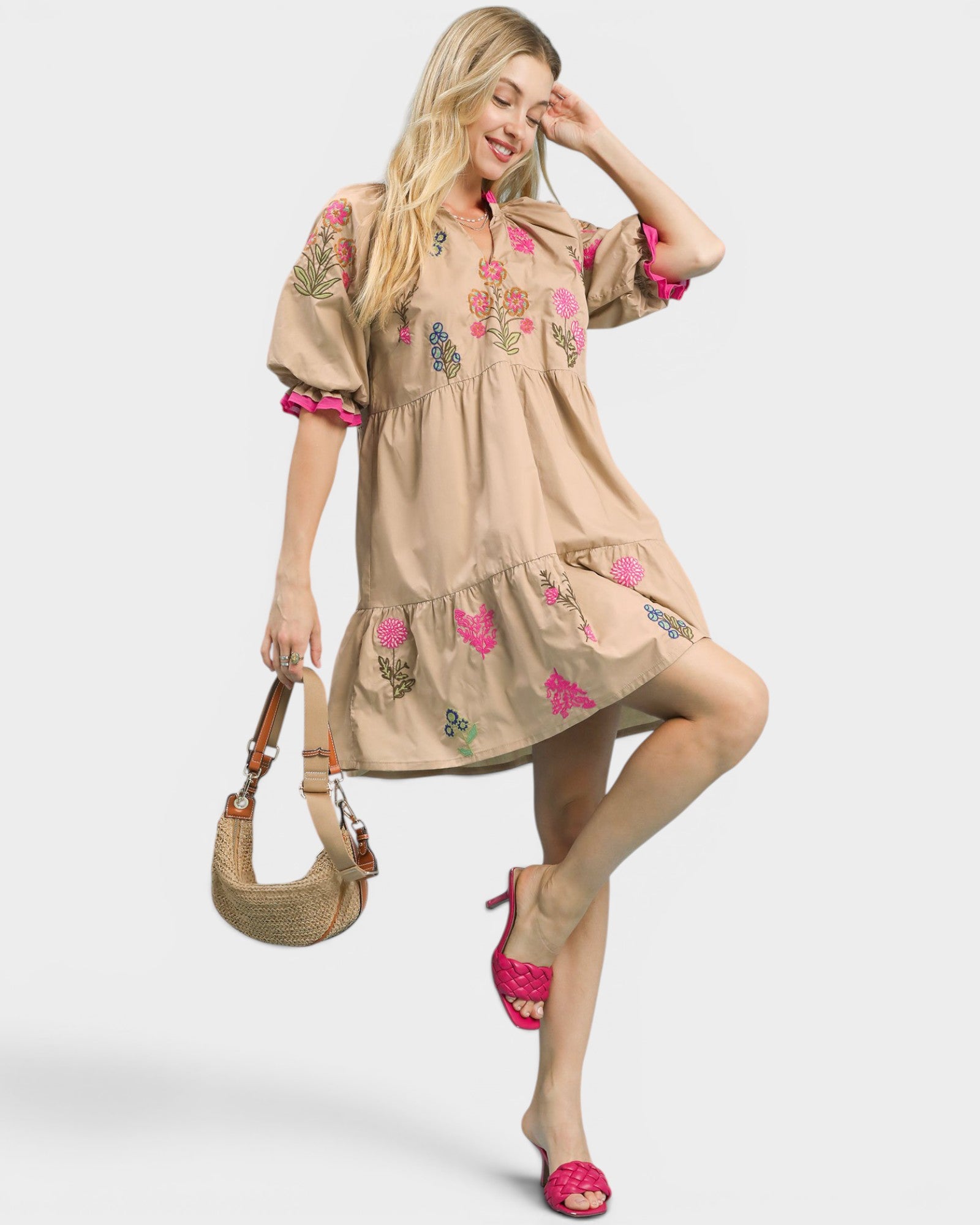 Woman wearing a beige dress with floral embroidery, holding a straw bag on a white background