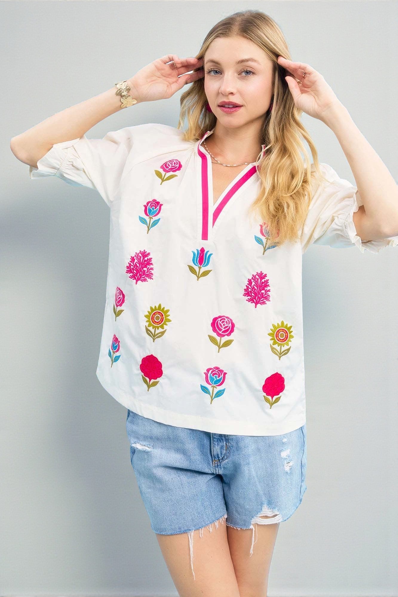 Woman wearing a white blouse with floral embroidery and denim shorts on a gray background