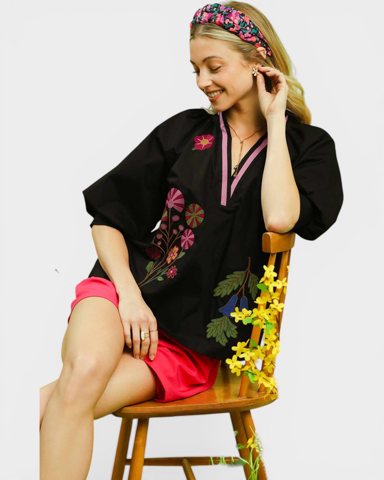 Woman wearing a black embroidered top and red shorts sitting on a wooden chair.