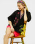 Woman wearing a black embroidered top and red shorts sitting on a wooden chair.