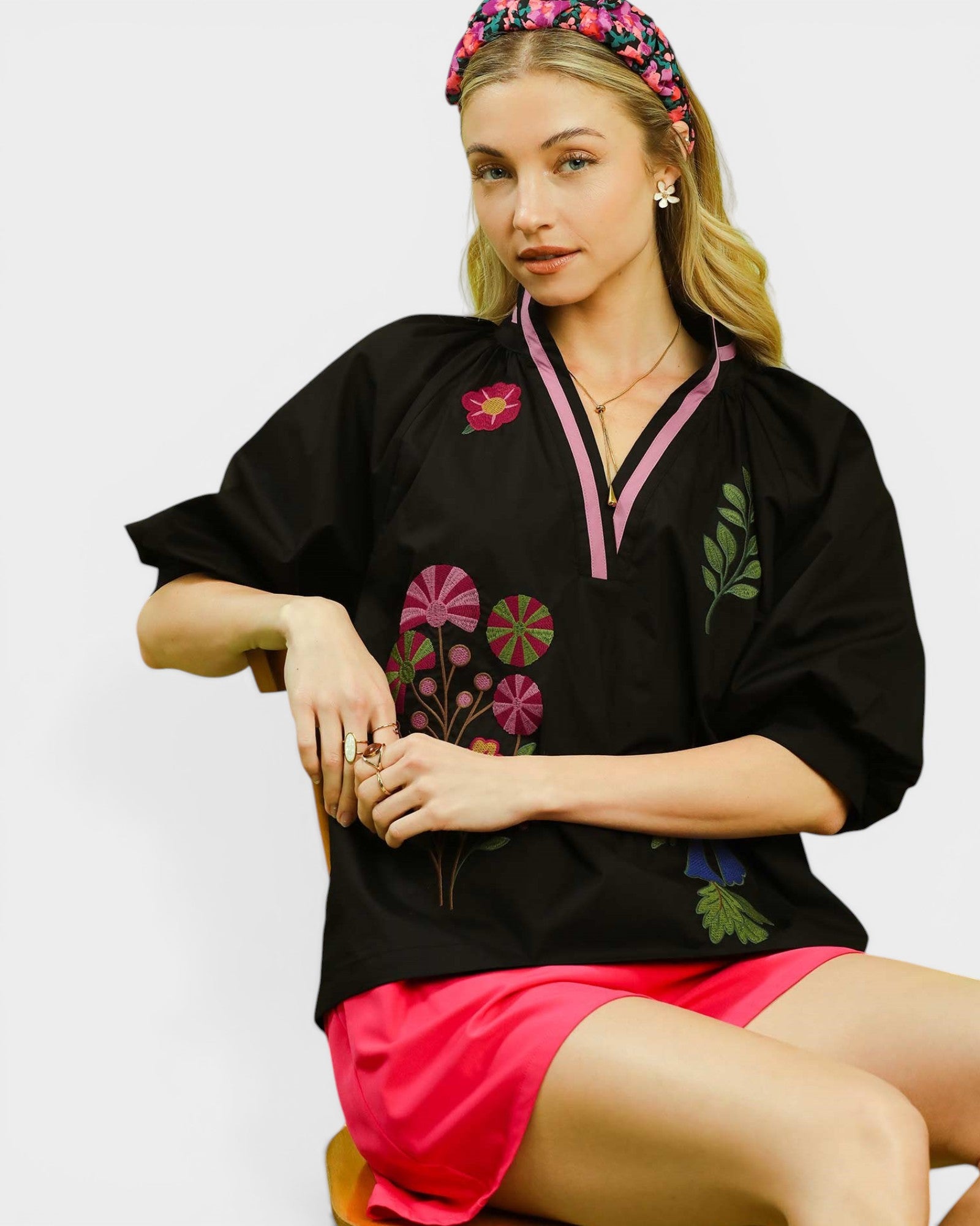 Woman wearing a black embroidered top and pink shorts with a floral headband on a white background