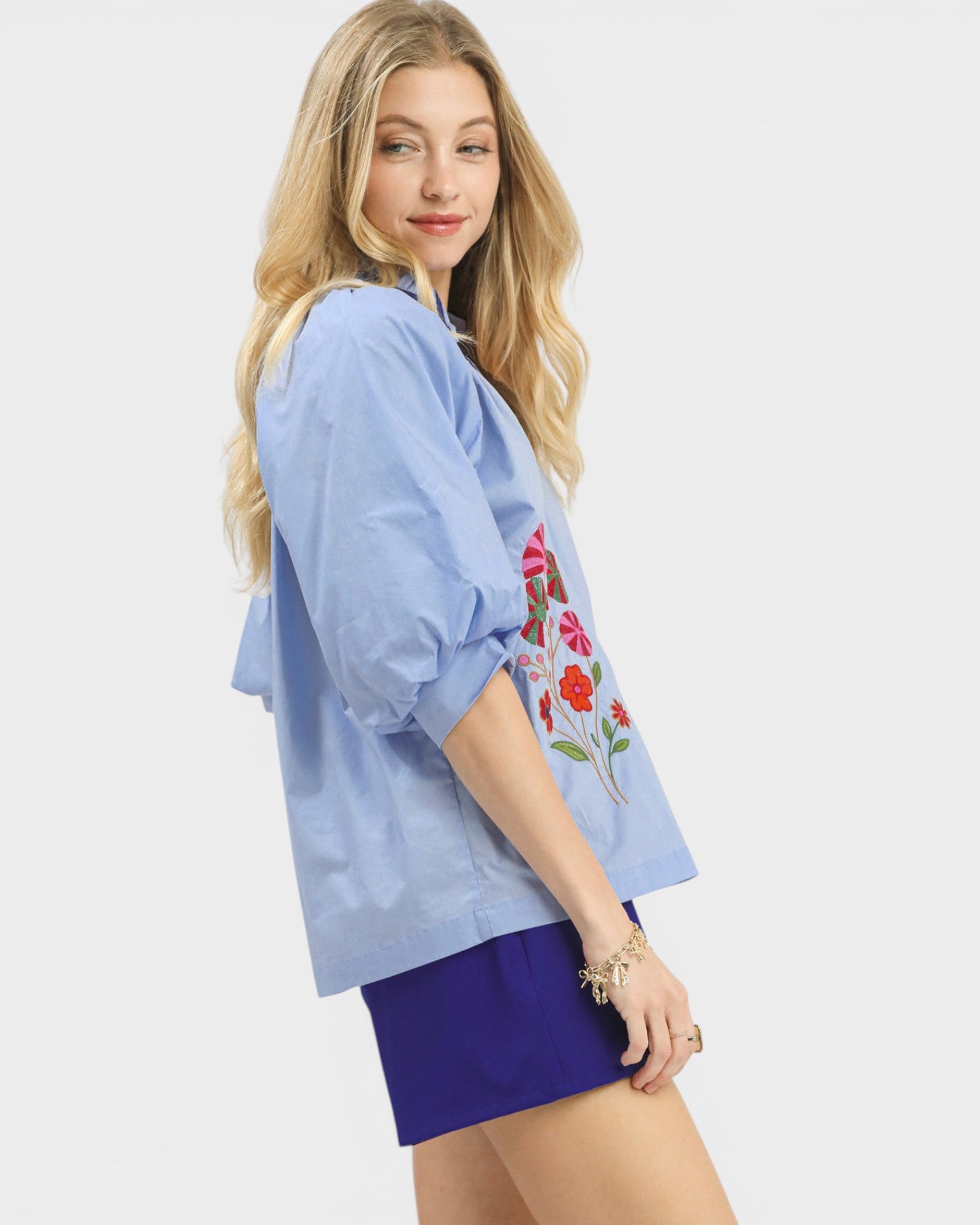 Woman wearing a light blue shirt with floral embroidery and purple shorts on a white background