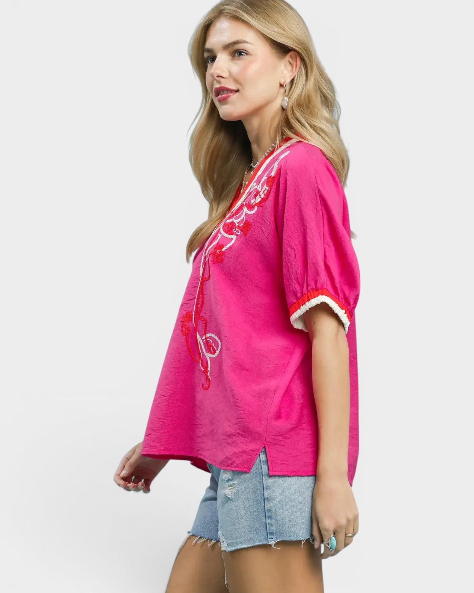 Woman wearing a bright pink embroidered top with denim shorts on a white background