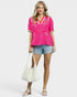 Woman wearing a pink embroidered top and denim shorts, holding a white bag on a white background