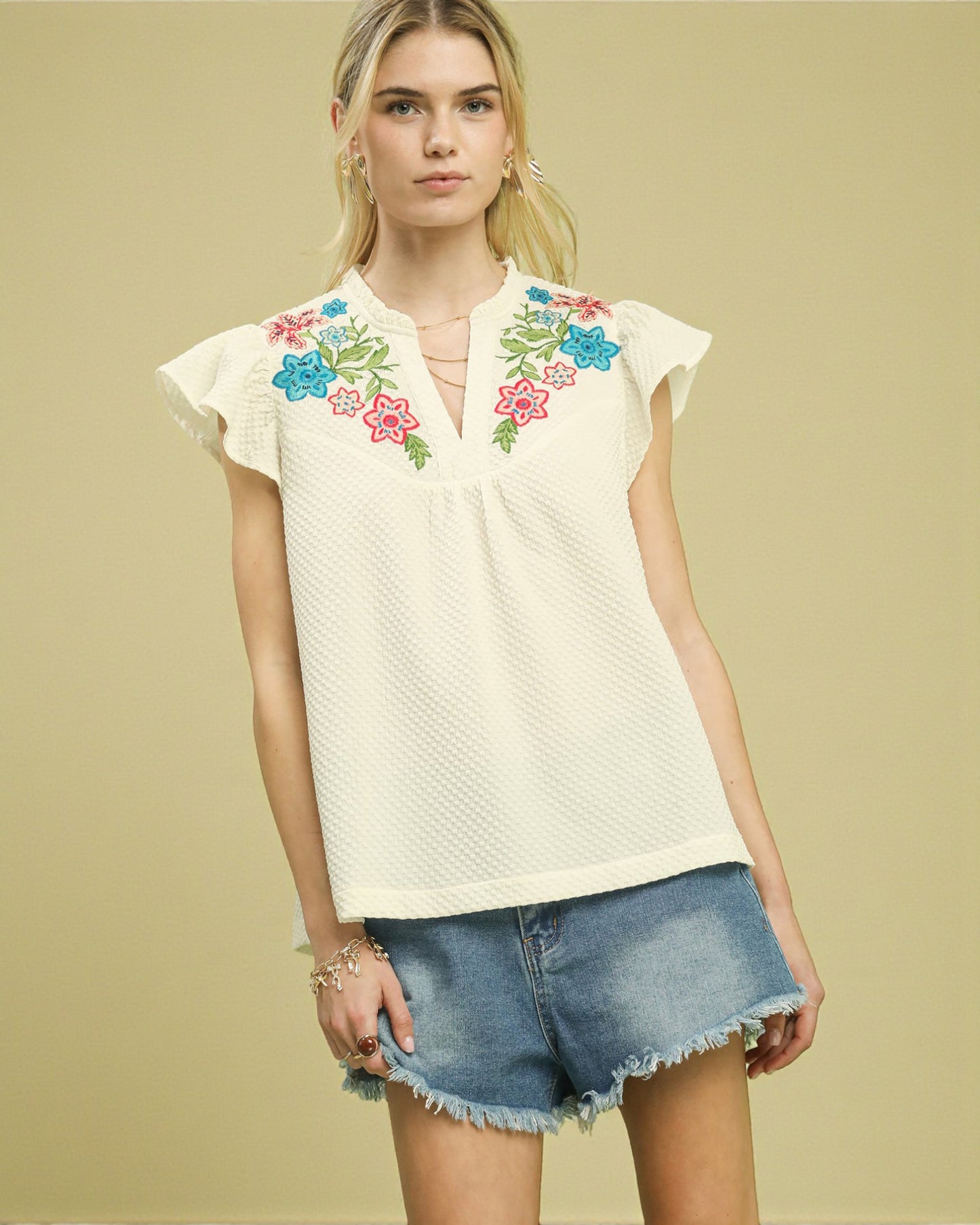 Woman wearing a white blouse with colorful floral embroidery and denim shorts on a beige background