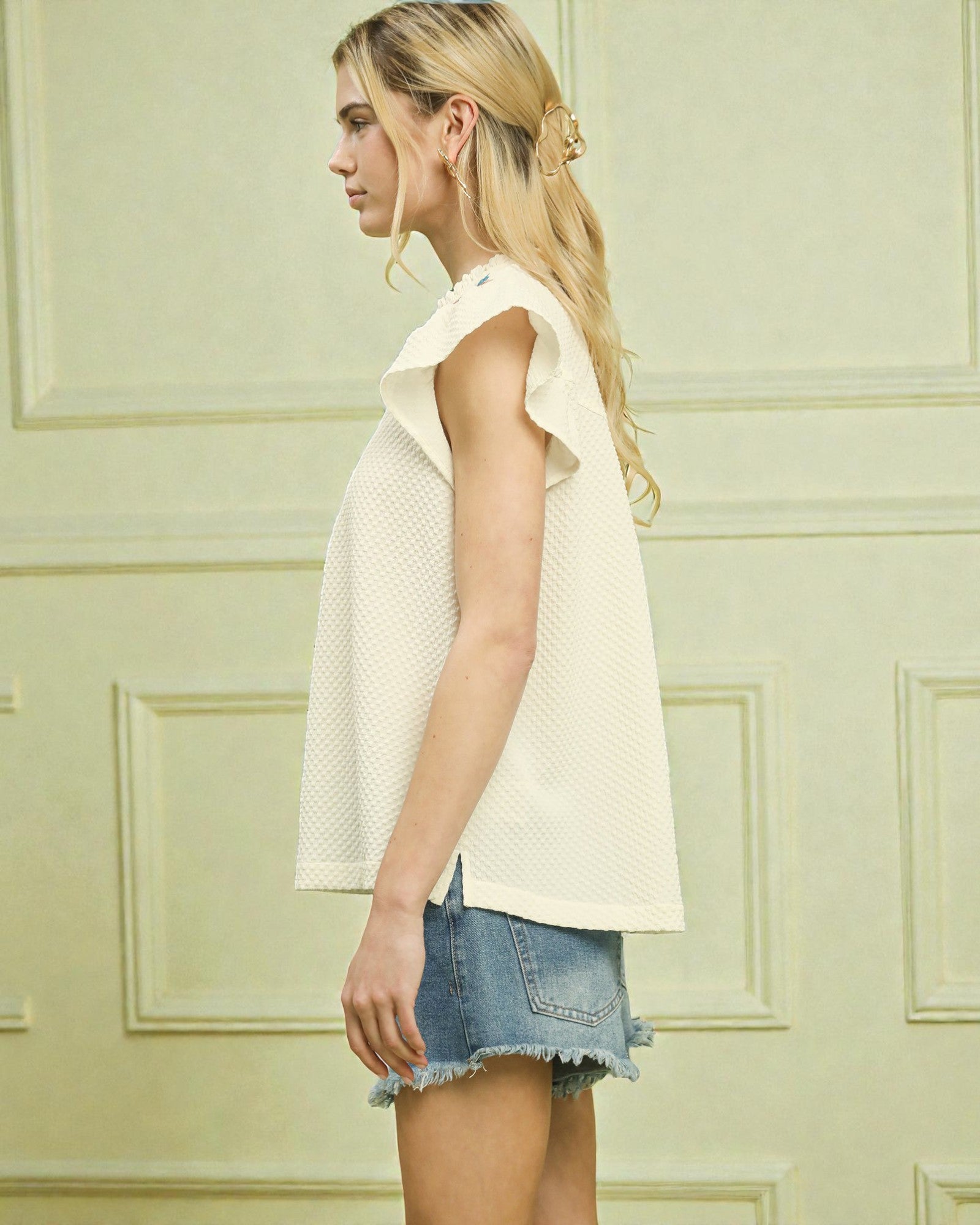 Woman wearing a light-colored sleeveless top and denim shorts against a light green wall.