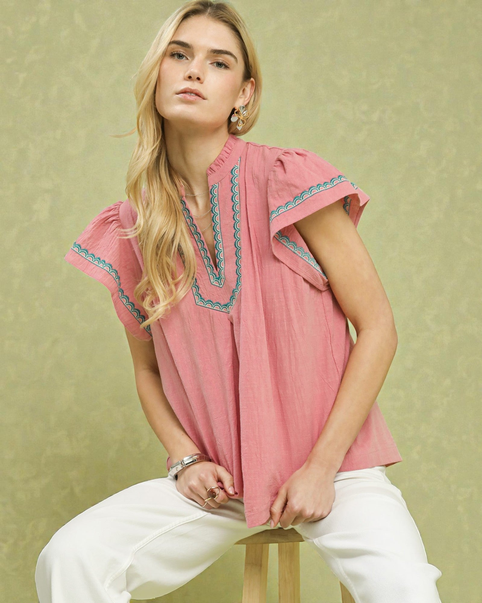 Woman wearing a pink blouse with embroidery sitting on a chair against a green background