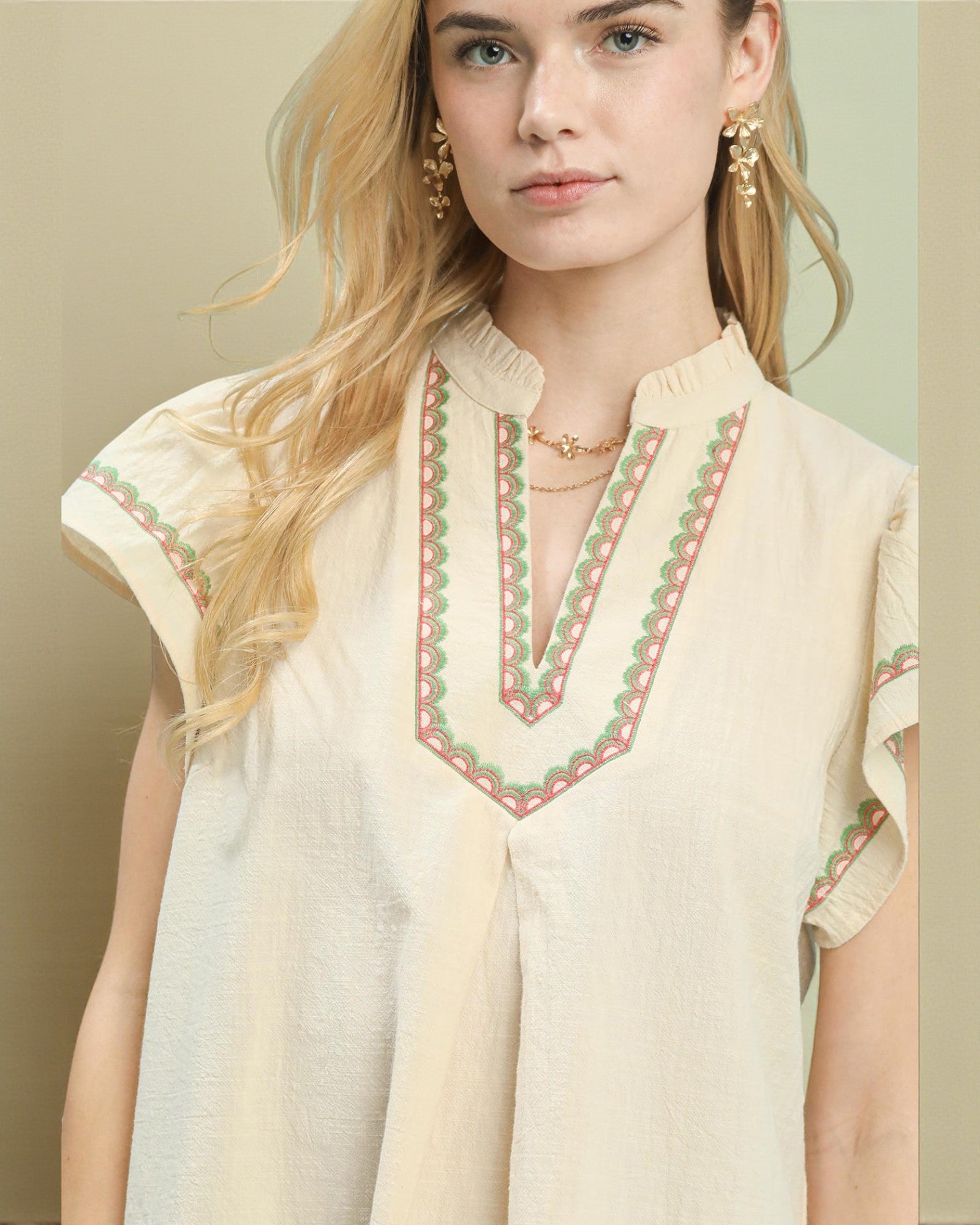 Woman wearing a cream-colored top with intricate green and pink embroidery on a neutral background