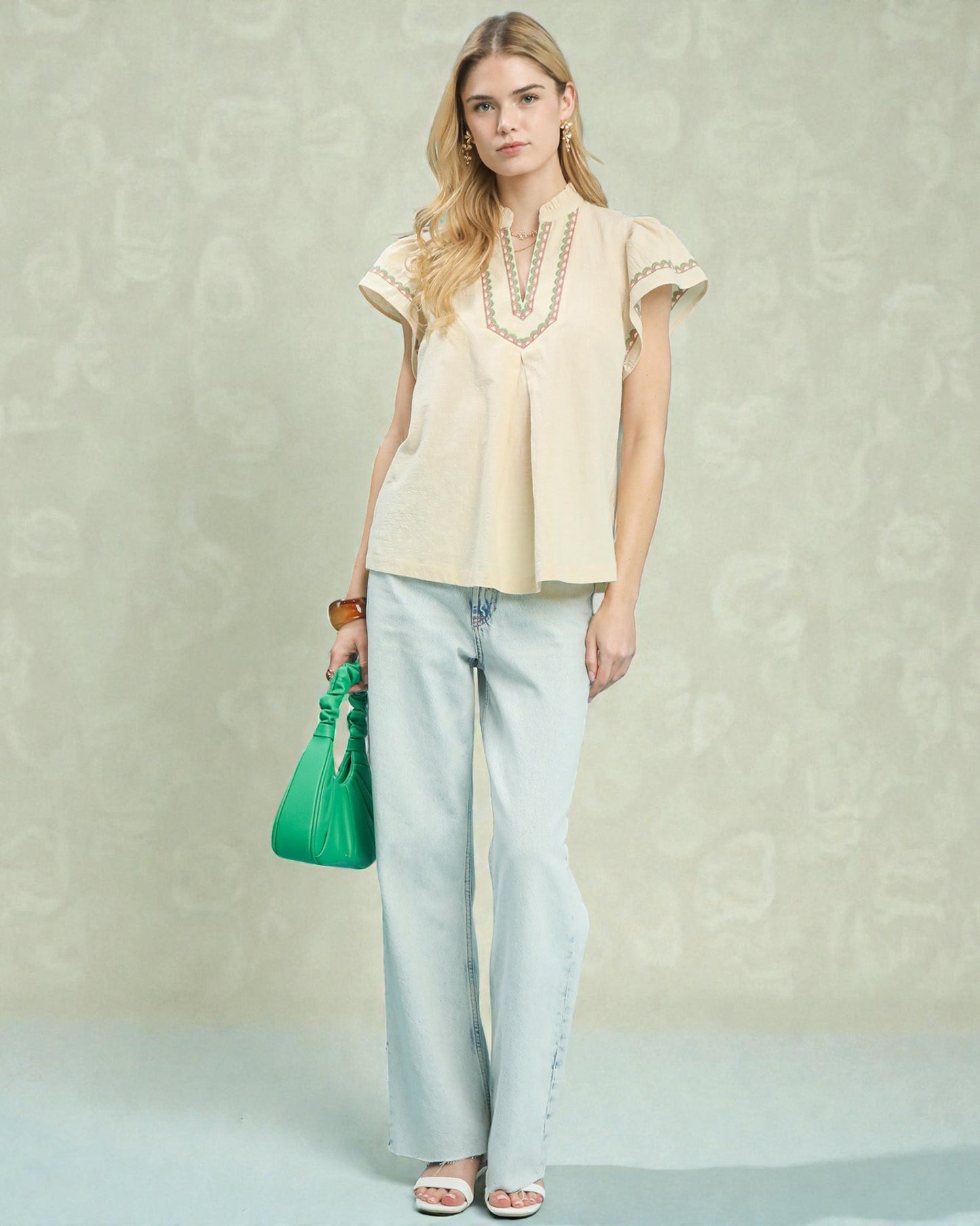 Woman wearing a beige blouse and light blue jeans, holding a green handbag against a neutral background