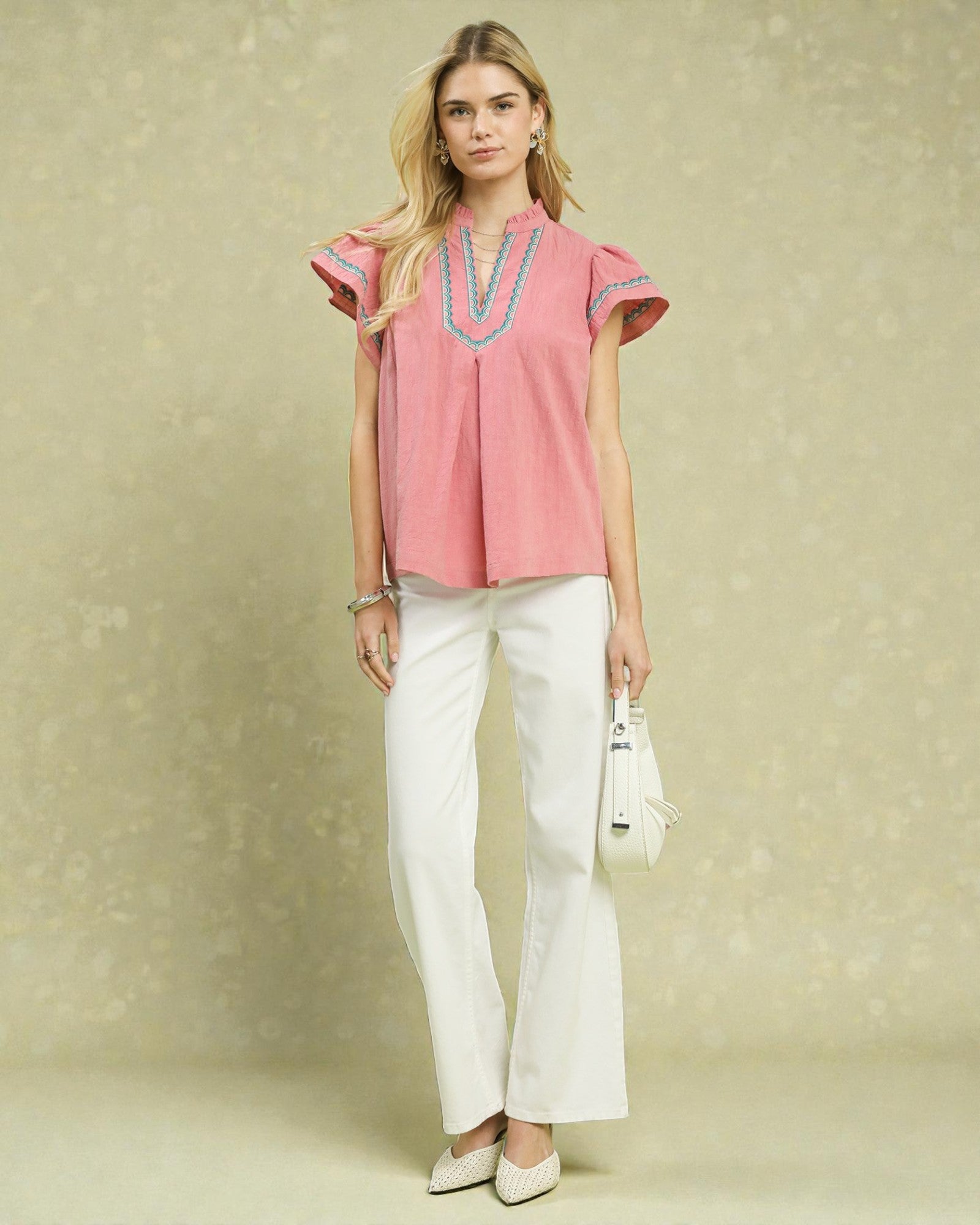 Woman wearing a pink top with embroidered details and white pants against a beige background