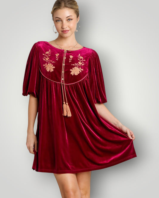 Woman wearing a burgundy velvet dress with floral embroidery on a gray background