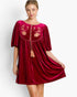 A woman models a short-sleeved burgundy velvet dress with gold floral embroidery and tassel ties on the chest, set against a plain light gray background.