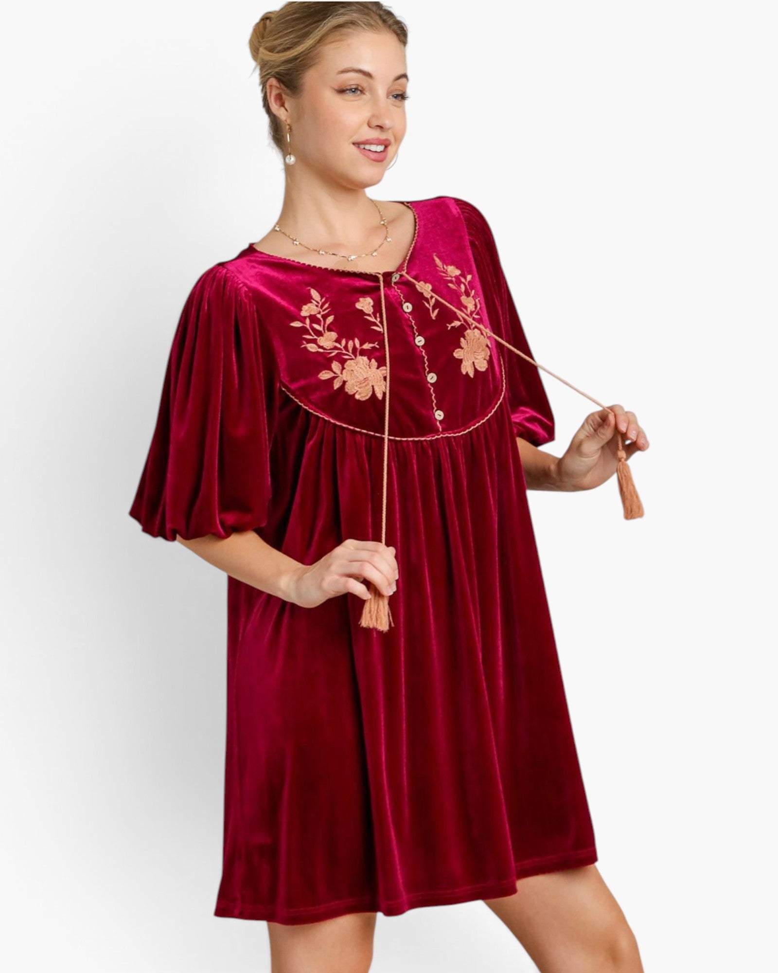 A woman modeling a short, loose-fitting velvet dress in deep burgundy red with gold floral embroidery across the chest and a buttoned neckline. She is holding a decorative rope accessory against a clean white studio background.
