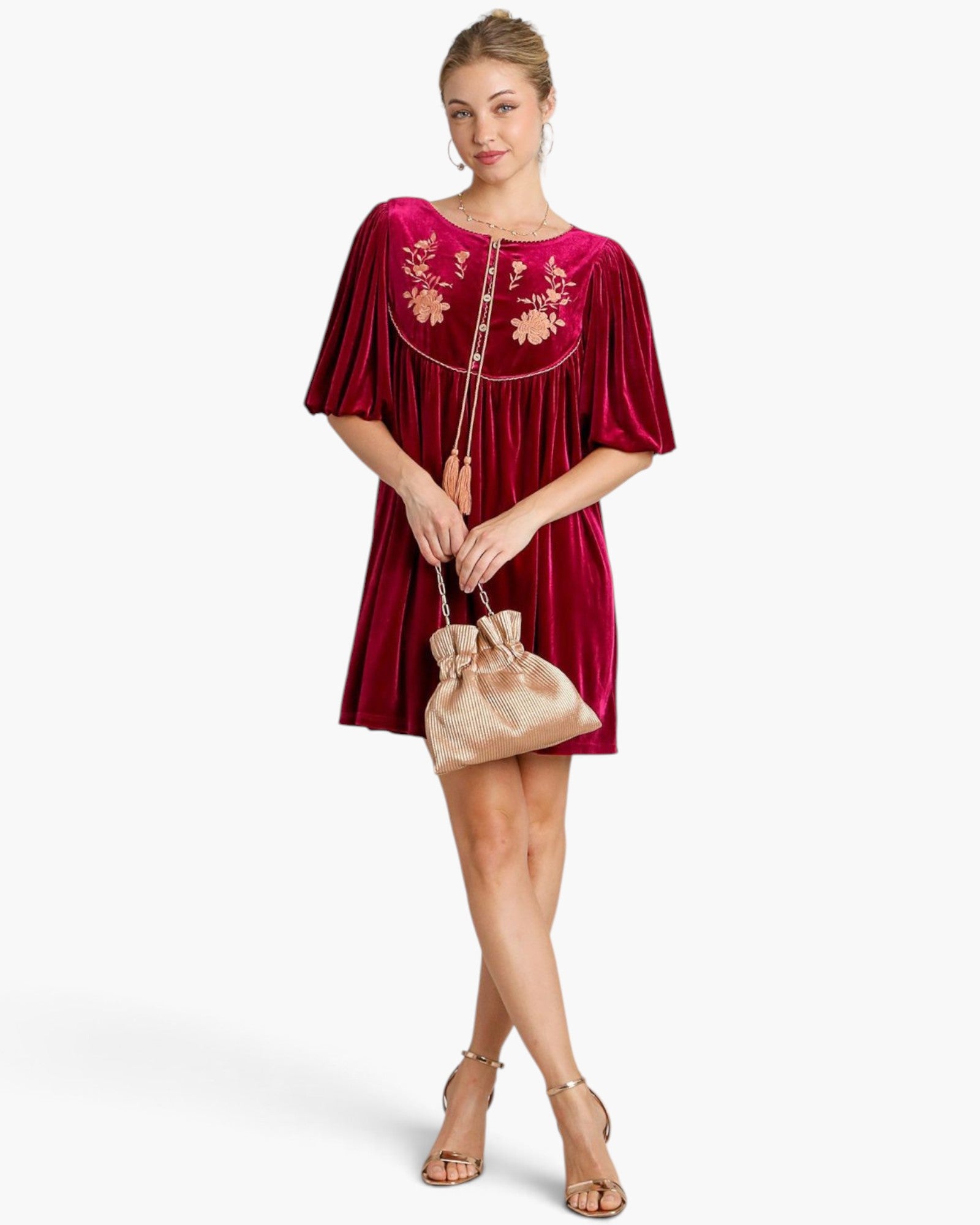 A woman models a short magenta velvet dress with floral embroidery on the chest and pleated short sleeves, holding a matching metallic pink drawstring pouch bag against a light gray studio background.