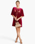 A woman models a short magenta velvet dress with floral embroidery on the chest and pleated short sleeves, holding a matching metallic pink drawstring pouch bag against a light gray studio background.