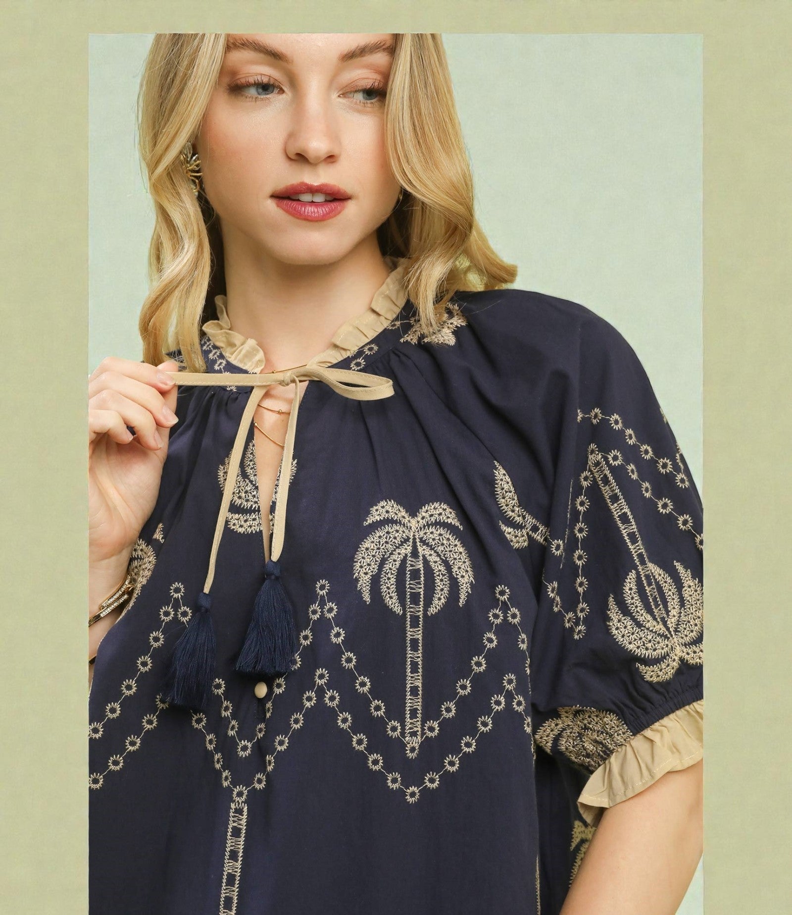 Woman wearing a navy blue blouse with gold embroidery and palm tree designs, holding a beige ribbon.