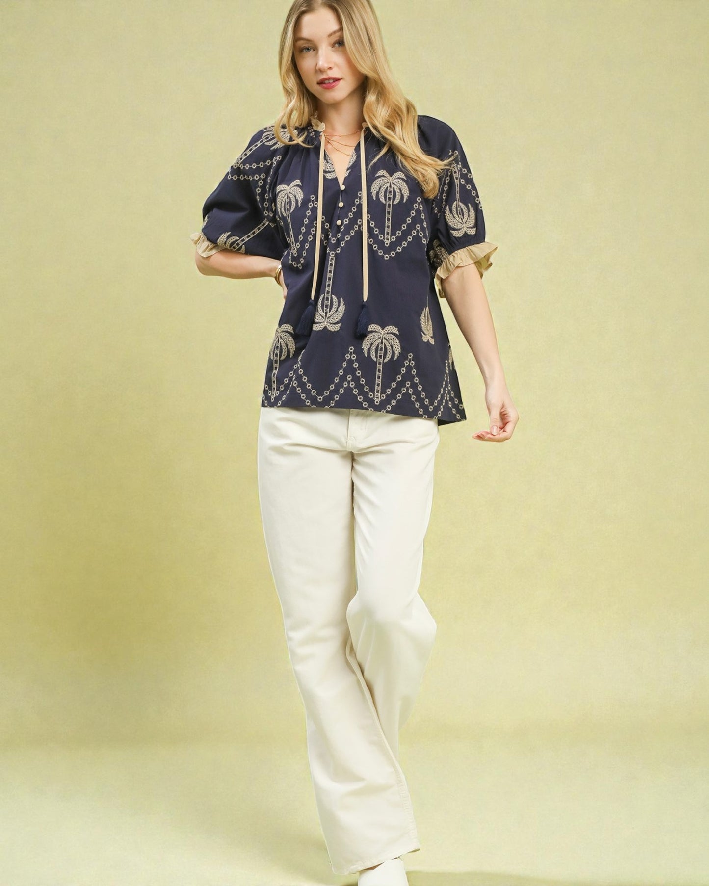 Woman wearing a navy blue embroidered top and white pants on a yellow background
