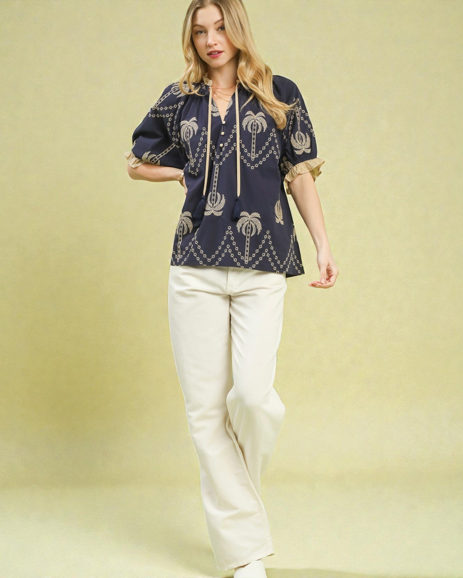 Woman wearing a navy blue embroidered top and white pants on a yellow background