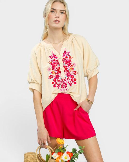 Woman wearing a beige top with floral embroidery and red shorts on a white background